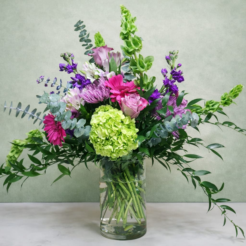 Majestic Harmony - Elevate any occasion with Majestic Harmony, a luxurious bouquet that blends the richness of purples and greens in an elegant display. This premium arrangement features lush purple stock, vibrant green hydrangea, and the velvety beauty of purple roses, creating a stunning foundation of color and texture. Dark pink gerbera add a playful pop, while the soft, ruffled petals of purple fujis bring a sense of sophistication. Lavender alstroemeria and delicate eucalyptus provide a soft, aromatic touch, with ruscus offering a refined, leafy contrast.    Displayed in a sleek glass vase, this arrangement exudes timeless beauty and grace, making it the perfect gift for someone truly special. Whether celebrating a milestone or simply expressing admiration, Majestic Harmony is an exquisite choice for those who appreciate the finer things in life.  approx 22" H x 22" W  PLEASE NOTE: Some flowers, vases and other products pictured are subject to availability, and may be substituted for a similar item if unavailable according to our substitution policy.