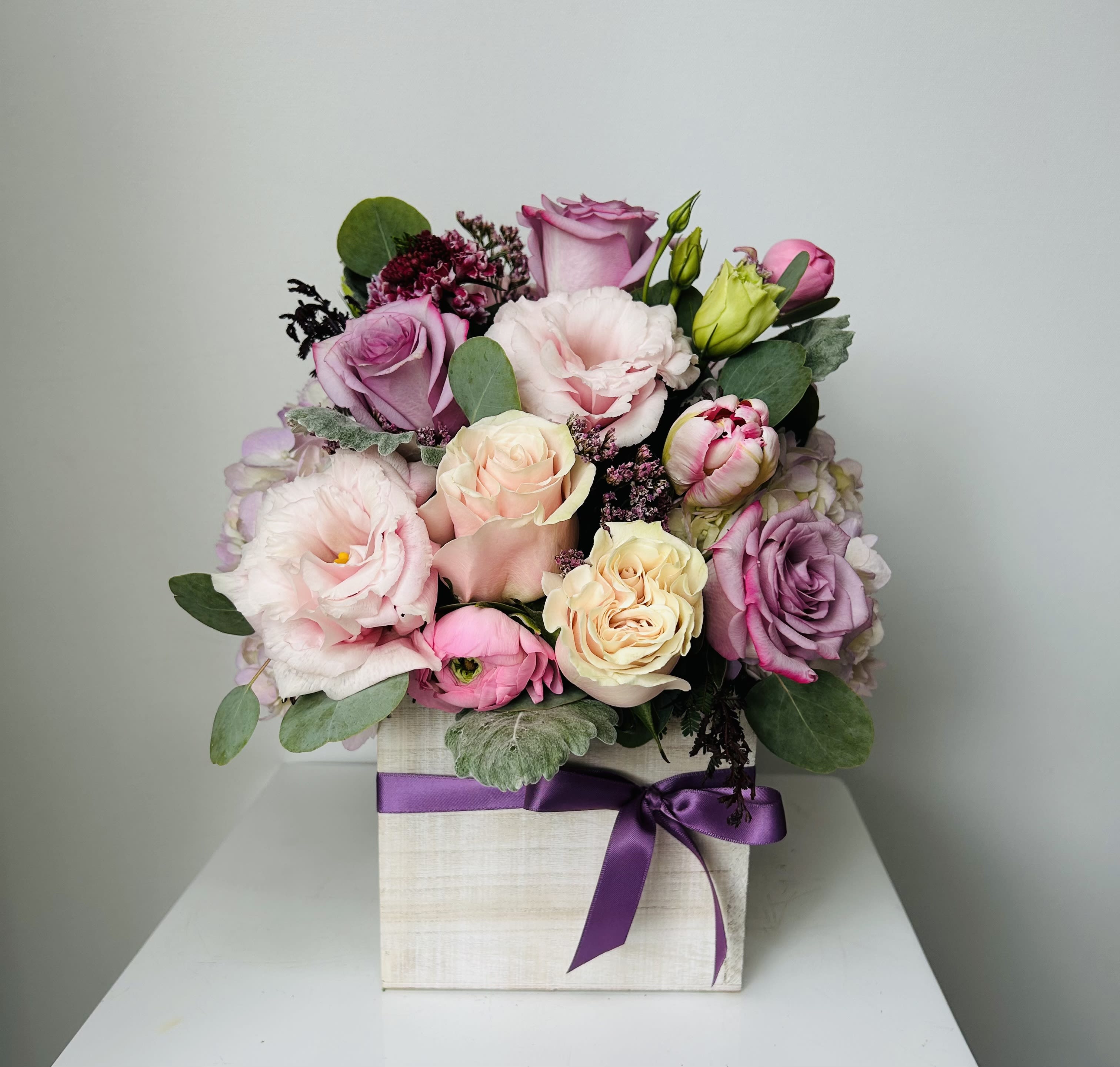Pink and lavender roses arranged in a square box with a purple ribbon