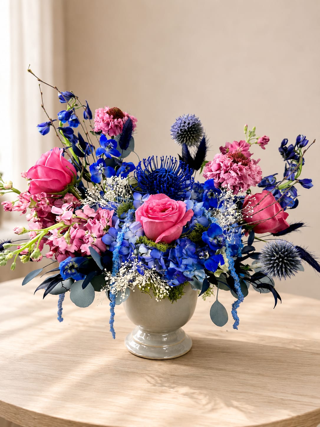 Emily  - Bright, lively color pops of vivid blue and soft pink make this Emily flower arrangement a joyful choice for a birthday bouquet, anniversary roses alternative, or Dia de los Muertos remembrance that still feels celebratory. The rounded ceramic vase or elegant white pedestal vase gives the design a polished, hand-crafted look from a San Antonio florist, and the dramatic, garden-style silhouette - stems and accent sprays reaching outward - ensures the person you're sending it to will feel genuinely seen.  Emily mixes pink roses and pink dianthus with clusters of blue hydrangea-like blooms, tall spiky delphinium or stock accents, creamy white filler daisies and airy branching stems for layered texture and contrast. Available in a compact low vase or a taller pedestal ceramic vase, the arrangement reads bright and romantic with a touch of modern drama; seasonal substitutions may be used to keep the same color palette and value while maintaining the overall style. 