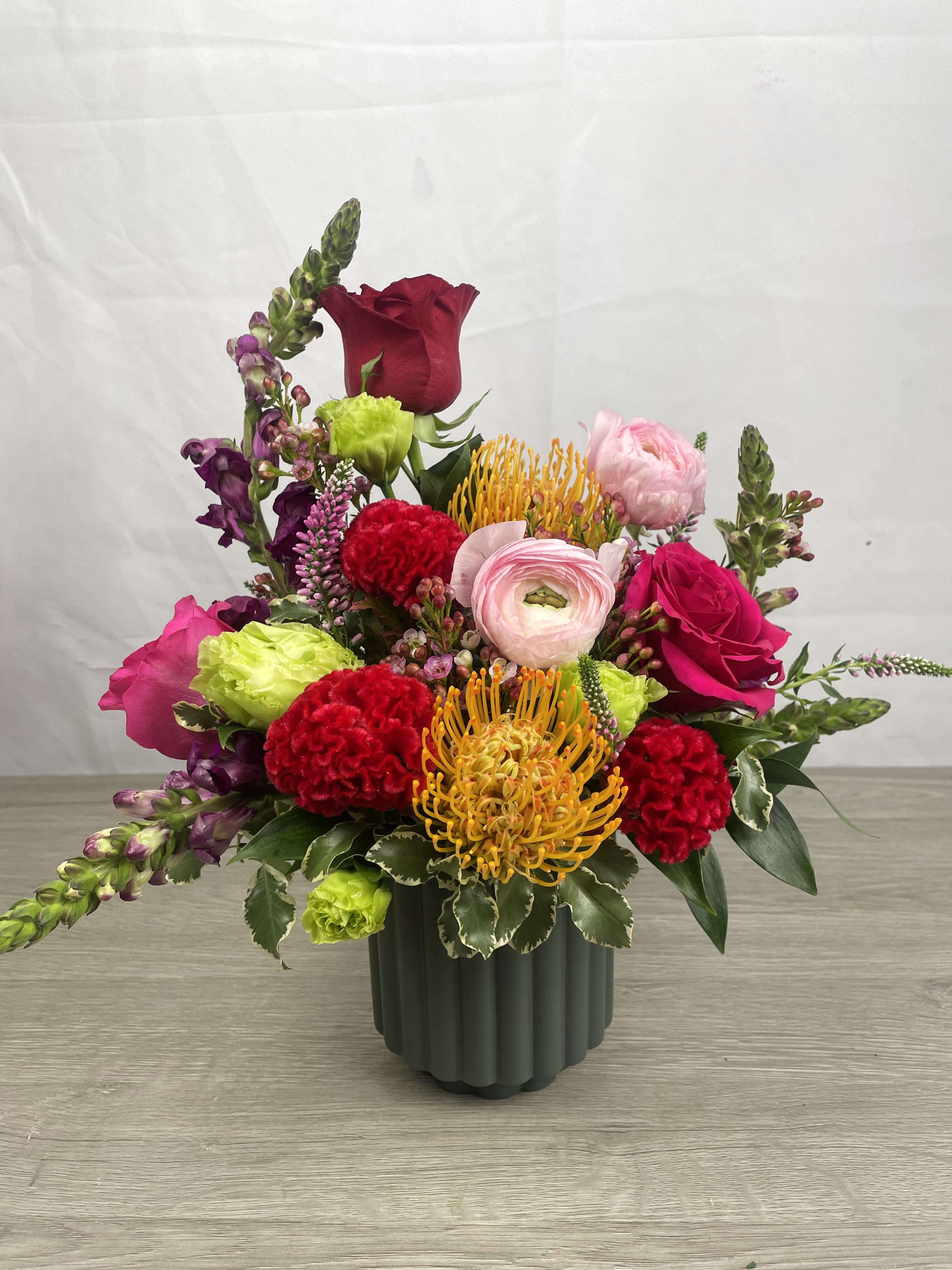 Velvet Fire  - Velvet Fire bursts into the room with a lively mix of red, pink, yellow and purple blooms-perfect as a birthday bouquet, anniversary arrangement, or a "just because" surprise. This mixed-color flower arrangement is designed to make the person you're thinking of feel noticed: romantic roses tuck into sunny ranunculus and cheerful carnations, while snapdragons and a protea-like focal bloom add height and a slightly wild, celebratory texture. As a San Antonio florist favorite, it reads as both intimate and joyful-ideal for sending love across neighborhoods from Alamo Heights to Stone Oak.  Tightly arranged in a dark green ribbed ceramic vase, Velvet Fire pairs rounded petals with spiky accents for a modern, hand-crafted look. The compact design fits tabletops and desks, and the vase gives the piece a polished, contemporary finish that suits downtown condos or suburban mantelpieces alike. Because we source fresh flowers in San Antonio, seasonal substitutions may occur to keep the same bold color palette and value while using the best, freshest stems available. 