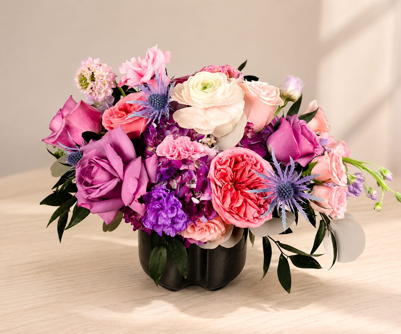 Velvet Blush - A lavish cluster of pink, lavender, coral and white blooms packed into a matte black vase-Velvet Blush reads like modern romance for an anniversary bouquet, birthday surprise, or just-because gift that makes the person you're thinking of feel truly seen. This rounded flower arrangement also dresses up get well wishes, new baby celebrations, graduations, Mother's Day, or a thank-you moment, and even carries a subtle Halloween vibe without getting spooky. As a San Antonio florist offering hand-delivered flowers in San Antonio, we style Velvet Blush to suit mantels in Stone Oak, coffee tables Downtown San Antonio, or bedside tables in Alamo Heights.  Inside the black vase you'll find lush roses and layered ranunculus-like blooms paired with lisianthus, stock, spray roses, carnations, dianthus and clustered accent flowers for a dense, textured look. Spiky blue thistle-like accents and darker foliage give a dramatic contrast to the soft pastels, creating a low, rounded centerpiece that reads like polished home décor. The design complements ceramic bowls, decorative books, woven planters or a potted plant and is sized perfectly for coffee tables and tabletops.  We may substitute seasonal varieties while keeping the same color palette and overall value so you always receive fresh flowers San Antonio can rely on. 