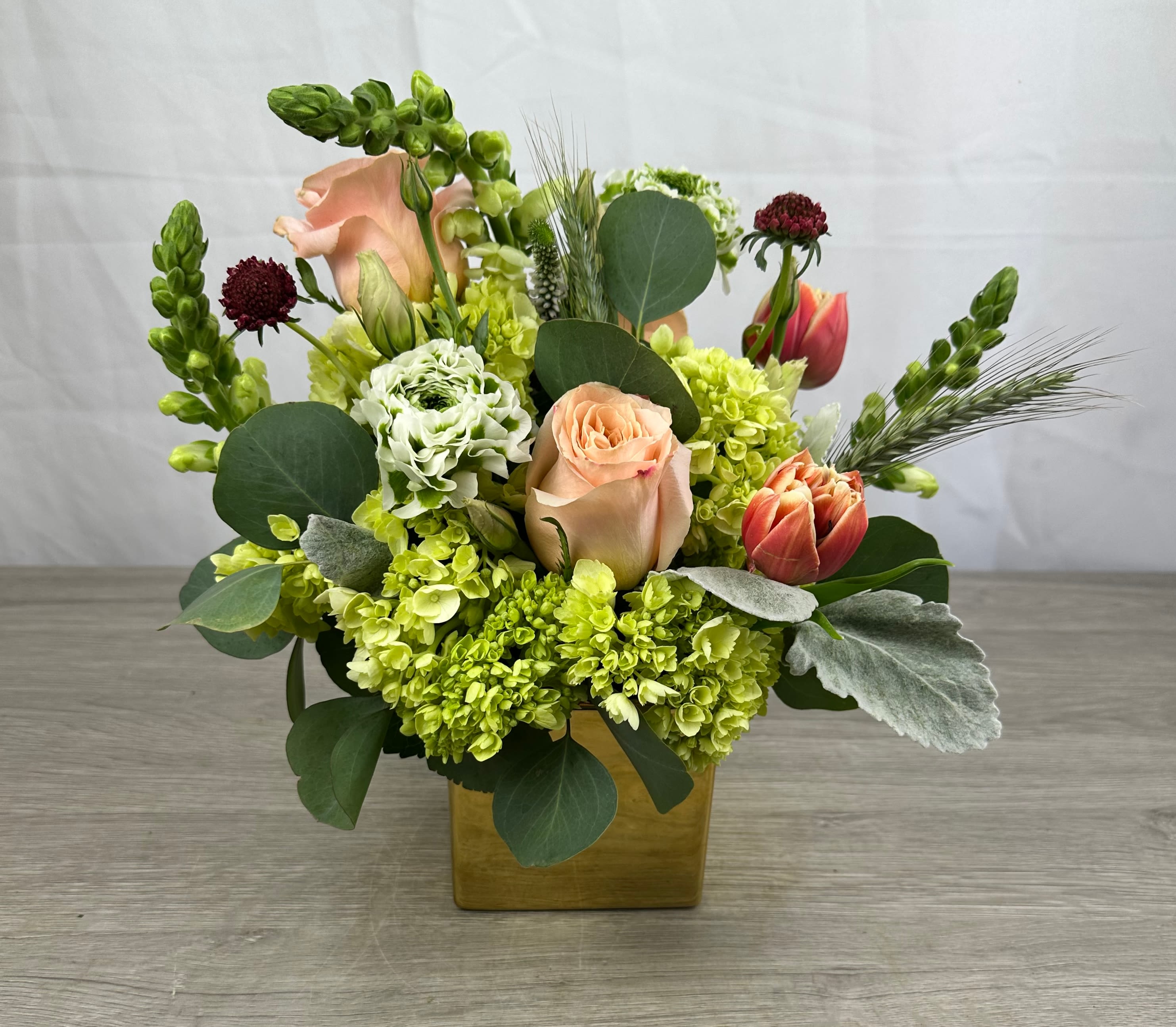 Goldie  - Soft peach roses and vivid chartreuse-green hydrangea clusters give this flower arrangement an instant fresh-spring lift-the perfect birthday bouquet or thank-you gift for someone who loves garden-fresh color. Designed in a compact square ceramic vase, Goldie pairs ruffled white blooms and pink-orange tulips with tall snapdragon spikes to create an uplifting, cheerful statement that still feels polished for desk or dining-table display.  Broad eucalyptus-like foliage and silvery dusty miller add layered texture while decorative grass and a few burgundy accents bring movement and contrast. The mix of roses, hydrangeas, tulips, snapdragons and other seasonal stems sits neatly in the low square vase for easy placement; it's the kind of arrangement that makes the person you're thinking of feel genuinely celebrated and brightens any spring celebration or everyday moment.