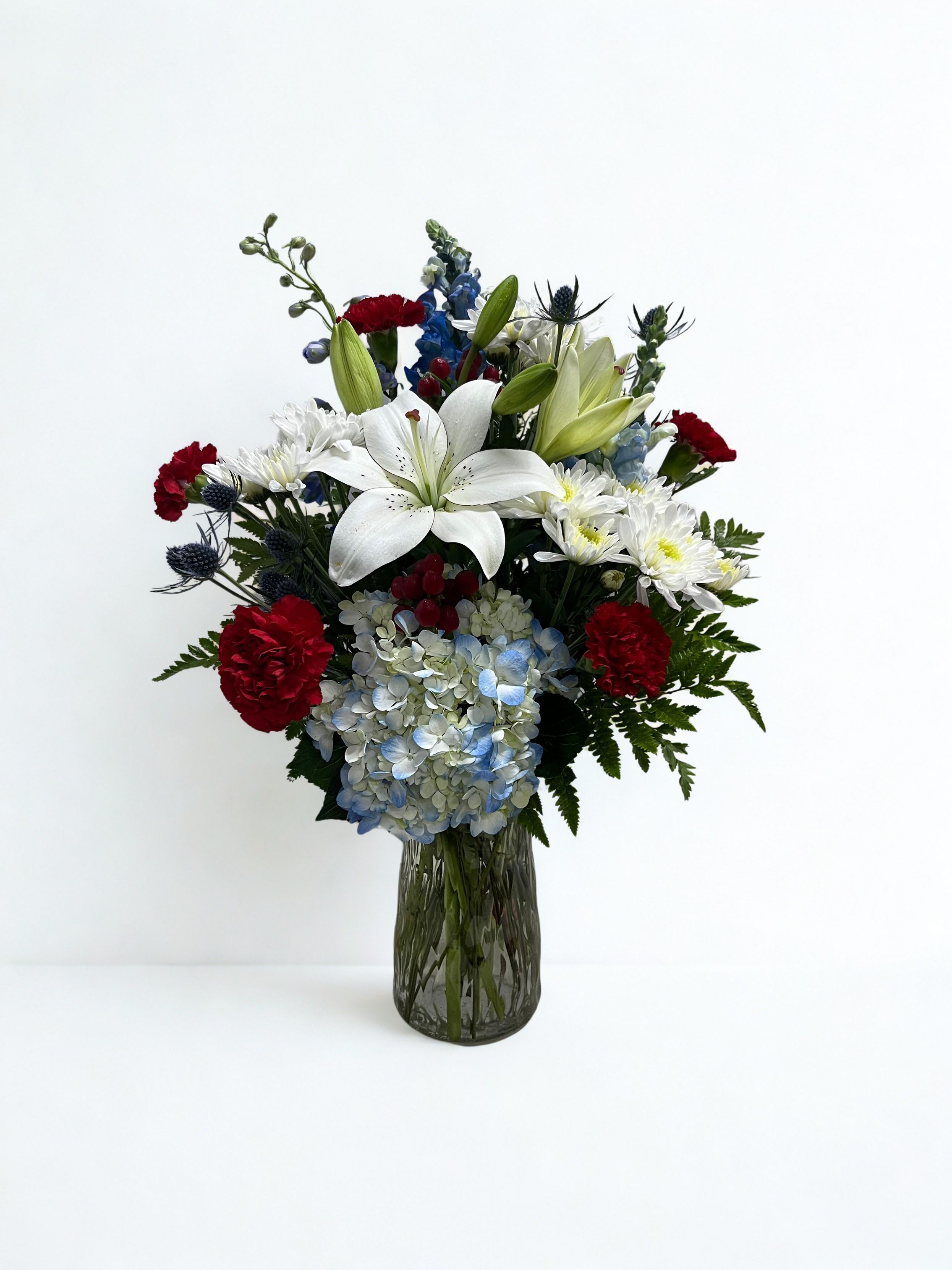 Freedoms Light - A single large white lily crowns this upright red, white, and blue flower arrangement-an arresting focal point that makes the bouquet feel both reverent and celebratory. Perfect as a sympathy or funeral tribute, a thoughtful Memorial Day or Fourth of July display, or a dignified thank-you or birthday gift for someone who loves classic patriotism, the arrangement reads as composed and respectful while still feeling bright and hopeful for happier occasions.  Rounded blue hydrangea clusters contrast with vivid red carnations, white daisy-like blooms, and tall blue snapdragon spikes to create layered texture and vertical interest. The palette of white, red, blue, and fresh green foliage is arranged in a structured, full form and comes presented in a dark glass vase that grounds the composition and suits formal settings.  Made to convey steadiness and care, this arrangement is the kind of gift that helps the recipient feel truly seen-whether you're honoring a life, saying thank you, or marking a birthday with quiet dignity. The sturdy vase and upright design make it easy to place on a mantel, table, or memorial site; simple water changes and trimming stems periodically will keep blooms looking their best.  As similar as possible. Designer may make appropriate substitutions based on flowers availability. 