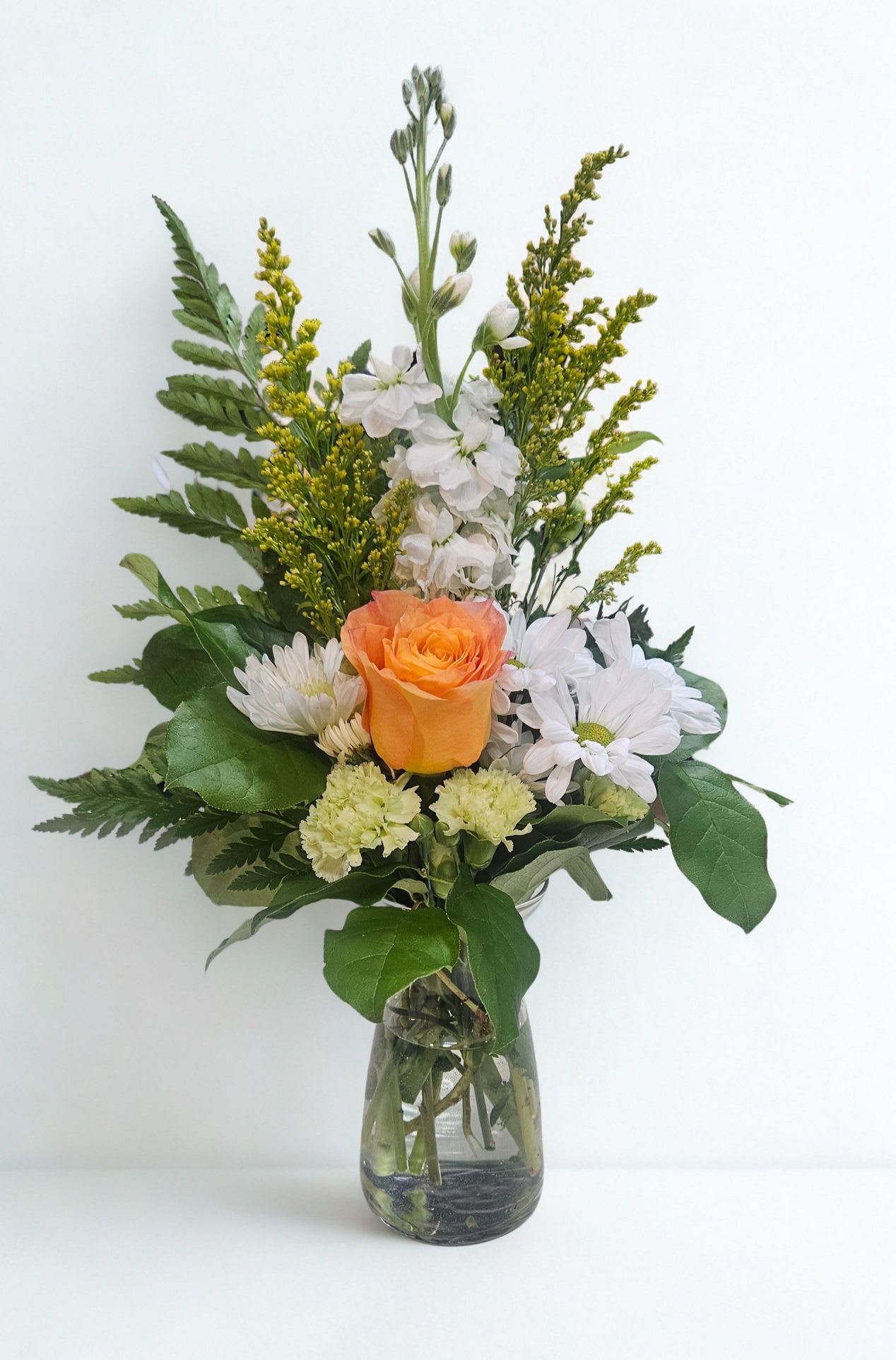 Fresh and Radiant - A single orange rose-warm, optimistic, and the immediate focal point-crowns this Fresh and Radiant flower arrangement, making it a joyful choice for a birthday, Mother's Day, or a heartfelt thank-you. The upright, loosely structured bouquet reads as casual elegance, the kind of everyday gift that says "I'm thinking of you" or "congratulations on your new home" without fuss.  Soft white daisy-style blooms and pale green carnations surround the rose, joined by airy yellow filler and broad green foliage that adds texture and a fresh, garden-picked feel. Arranged in a clear glass vase, the design stays bright and uncomplicated so the colors-orange, white, yellow, and green-take center stage and brighten any room.  Perfect for sending just because or to mark a special moment like a birthday or housewarming, this arrangement feels equally at home on a kitchen table or a bedside dresser. The simple vase and sturdy stems make care easy: keep fresh water in the glass, trim stems occasionally, and display away from direct heat to extend the cheerful life of the blooms.