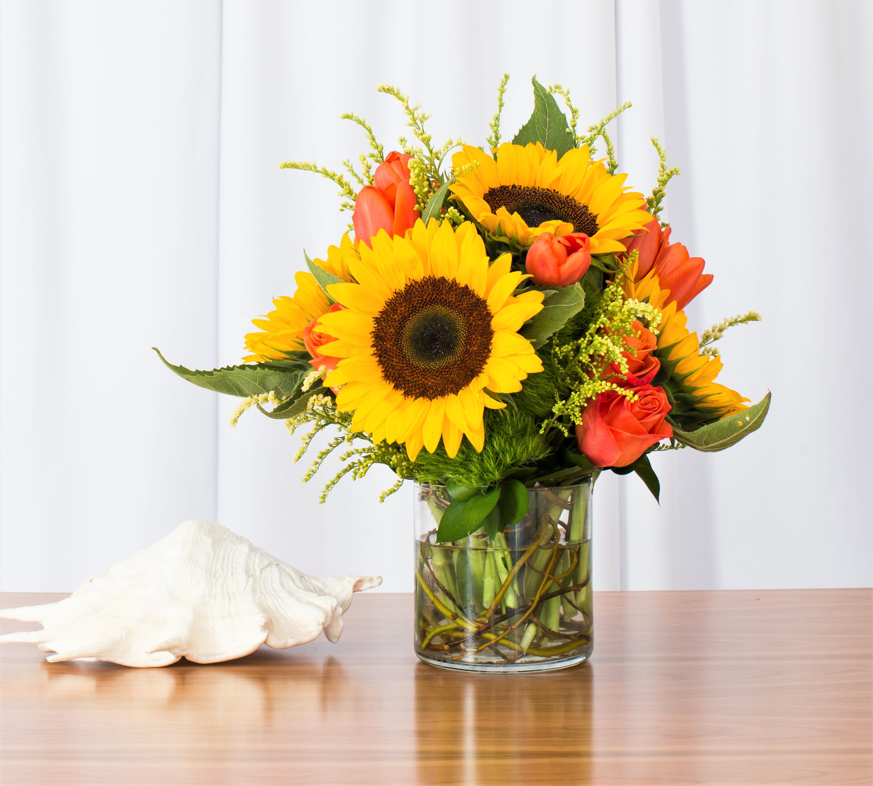 Yellow sunflowers and orange roses in a clear glass vase on a table with a white seashell.