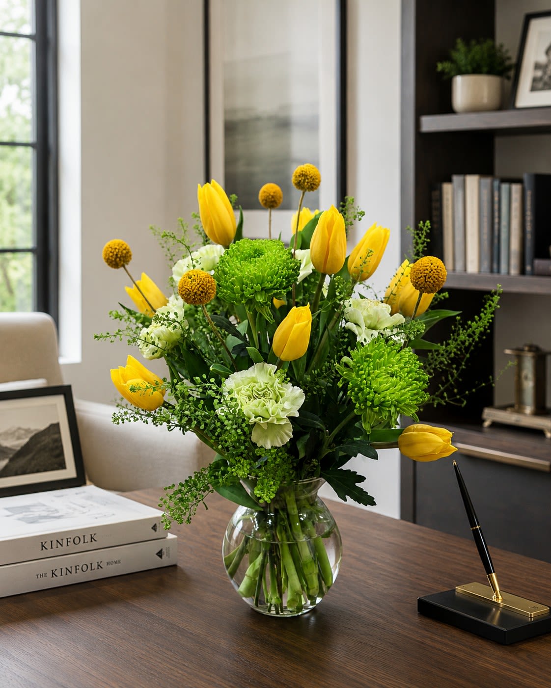 NEW! Citrus Twist  - Bright yellow tulips take center stage in this spring-ready flower arrangement, perfect as a birthday bouquet, cheerful thank-you gift, or everyday pick-me-up. The combination of tulips, pale green-white carnations, and vivid green chrysanthemums creates an uplifting yellow, green, and white palette that reads fresh and modern - the kind of arrangement that makes the person you're thinking of feel bright and noticed.  Arranged in a round clear glass vase, the design adds playful height with small spherical yellow pom‑pom blooms and a light, airy filler for a loose, garden-style texture. Compact yet lively, the bouquet blends smooth tulip petals, ruffled carnation texture, and daisy-like chrysanthemum form for visual contrast and lasting appeal.  Ideal for spring occasions, cheerful celebrations, or adding sunny color to a home or office. For freshness and best care, please refer to the Care and handling QR code.