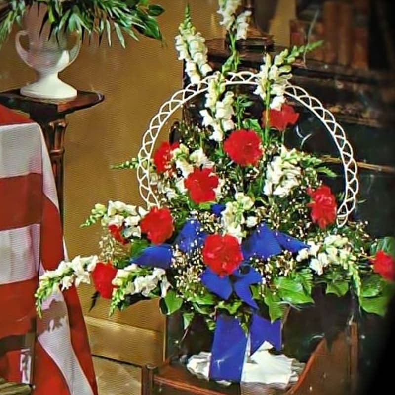 Patriotic red carnation and white snapdragon sympathy arrangement with blue bow in a container.