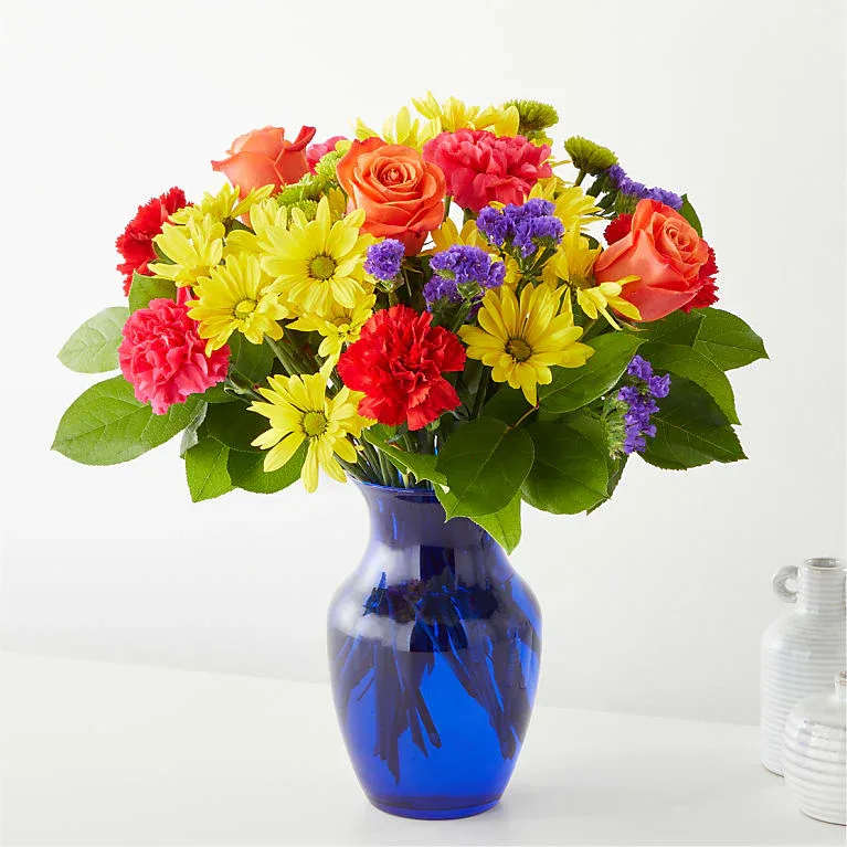 Bright &amp; Beautiful - Bright, cheerful, and full of personality, this lush rounded arrangement features sunny yellow daisies, vibrant orange roses, soft pink carnations, and rich purple accent blooms gathered in a charming blue glass vase. The lively mix of colors creates a joyful display that instantly brightens any room, making it a perfect gift for birthdays, celebrations, thank-you gestures, or simply to share a smile.
