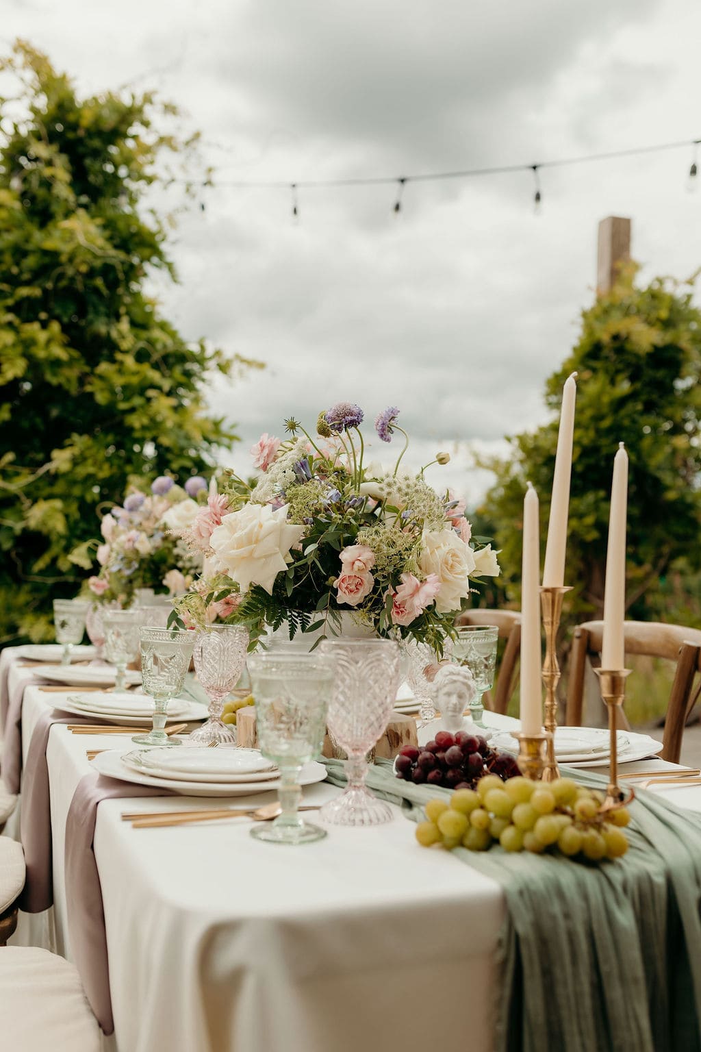 Pastel rose centerpiece on an outdoor banquet table with crystal goblets, taper candles, and grapes on a green runner.