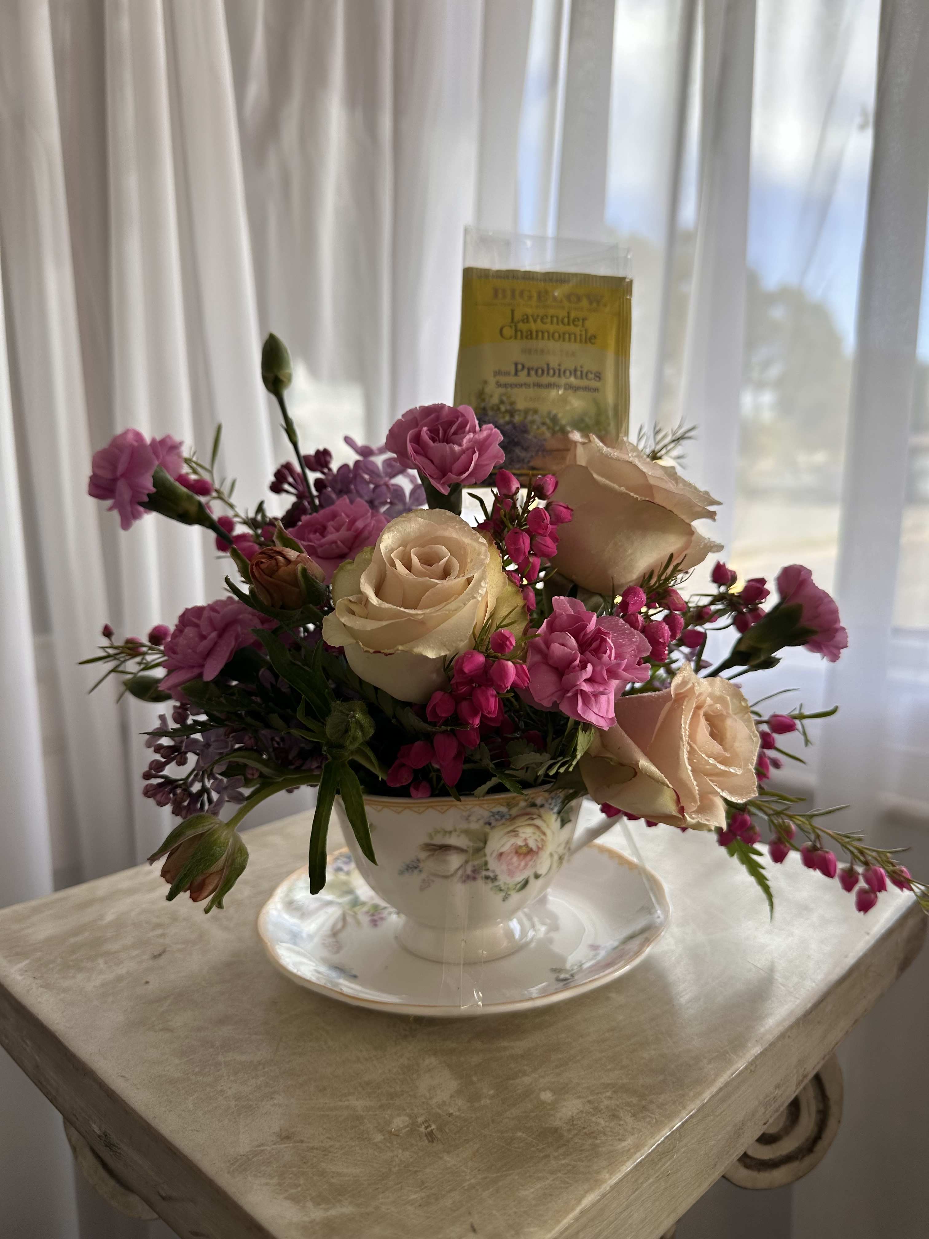 Pink and cream roses arranged in a teacup with a tea packet behind them.