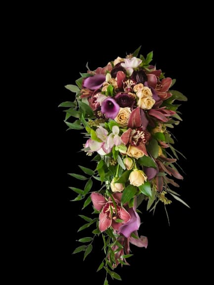 Tender Vows - A dramatic cascading bouquet designed for a wedding or special event, this arrangement makes the kind of impression that will leave the person you're thinking of speechless. The elongated silhouette and long trailing stem line give it a formal, romantic presence-perfect as a bridal cascade or a statement piece for anniversaries when you want the recipient to feel deeply celebrated.  Rich purple mini calla lilies sweep through the dense top, paired with cream roses and burgundy orchid blooms for a layered, textured look. Lush green foliage trails beneath and between the flowers to emphasize the bouquet's elegant drop; the mix of deep purples, soft pink-cream roses, and touches of white-green foliage creates a sumptuous, tactile color story that reads both classic and modern.  For a bride or honoree who loves dramatic florals, this bouquet signals romance and refinement while still feeling fresh. Its elongated shape suits formal ceremonies and photographs-handle with care and present it supported to preserve the trailing stems; consult our sizing options at checkout to choose the scale that best fits your wedding or event.