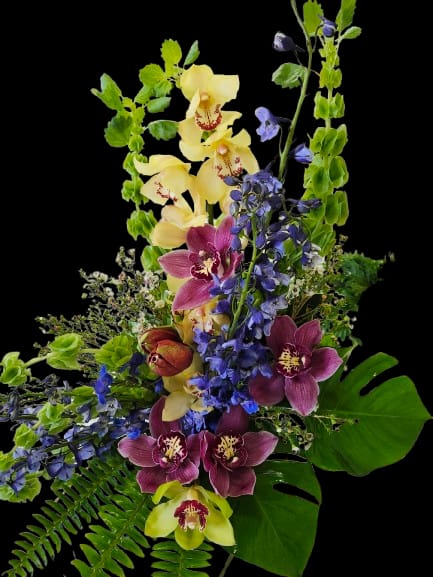Vibrant Joy - A tall, dramatic tropical-style flower arrangement that greets the room with bold upward lines and saturated color-perfect as a birthday bouquet, congratulations arrangement, or a cheerful everyday gift. Orchids in yellow, magenta and lime take center stage to create a vivid, celebratory impression while vertical sprays of blue delphinium add airy height and movement that catch the eye.  Lush tropical leaves and fern fronds form a textured, garden-like base that grounds the composition and enhances the orchids' exotic glow. The mix of purple, yellow and green tones gives the bouquet a playful, modern look that reads as joyful rather than delicate.  Choose this arrangement when you want someone to feel noticed and celebrated-ideal for a friend's milestone, a bright pick-me-up, or sending sunny wishes. Designed to stand tall and make an impact, it arrives ready to display; keep in fresh water and trim stems every few days to maintain peak freshness.