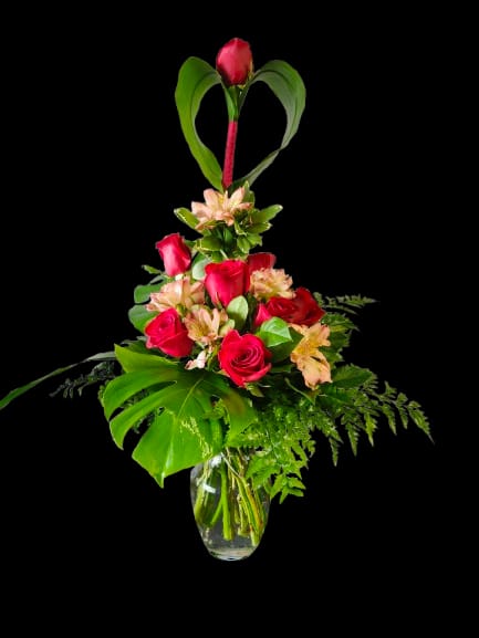 Heart in Bloom - Vivid red roses clustered with soft peach-pink alstroemeria create a romantic focal point in this flower arrangement, perfect as an anniversary bouquet or a heartfelt romantic gift. The tall heart-shaped leaf accent rising above the blooms adds a decorative, slightly whimsical touch that draws the eye and makes the recipient feel truly noticed.  Arranged in a clear glass vase, the mix of red, pink, white accents and fresh green foliage reads lush and celebratory. The combination of velvety roses and delicate alstroemeria offers both bold color and airy texture, making it equally at home on a dinner table or bedside. Sized for easy display, it arrives ready to enjoy-simply add water and trim stems every few days to keep the arrangement fresh.