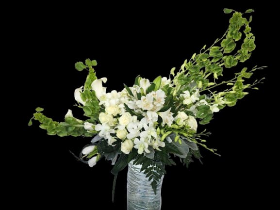 Airy Elegance - A tall, sculptural arrangement of white blooms with fresh green accents makes a striking statement for an anniversary, sympathy tribute, or elegant birthday bouquet. The soaring arching stems and layered textures create a dramatic yet soft presence that will make the person you're thinking of feel deeply noticed and comforted-perfect for an entryway, reception table, or any formal gifting moment.  Calla lilies, white roses, orchids, snapdragons and mixed-color alstroemeria are arranged with lush greenery in a tall glass vase wrapped with decorative wrap, giving the design height and airy movement. The palette reads serene and refined: pure whites and crisp greens that emphasize the flowers' sculptural lines and delicate textures. Sized to stand proudly as a focal piece, this arrangement arrives ready to display; trim stems and refresh the water every few days to keep blooms looking their best.
