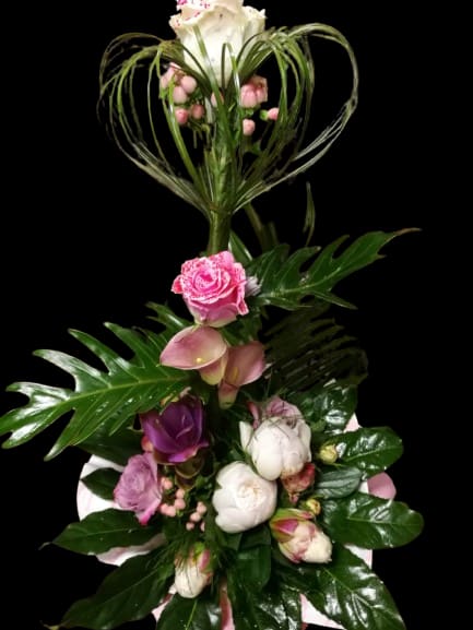 Tender Bloom - A dramatic vertical arrangement of pink and white roses and calla lilies creates an immediate romantic impression-perfect as an anniversary bouquet, birthday floral gift, or a "just because" surprise for someone you adore. The tall, sculptural line of blooms draws the eye upward, while the decorative heart-shaped loop of glossy greenery at the top makes the recipient feel truly seen and celebrated.  This design pairs soft pastel roses and calla lilies in pink and white with small pink filler blooms for texture and rich tropical foliage for contrast. The arrangement is intentionally tall and sculptural, with glossy leaves framing the flowers and a distinctive heart-shaped greenery accent that adds visual height and elegance. It's a refined, emotional statement bouquet that reads as both celebratory and intimate-ideal for sending warm affection or marking a special day.