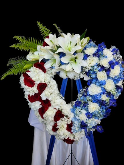 American Heart - A large heart-shaped sympathy arrangement that reads with quiet dignity: white lilies crown the top while clusters of red roses form the left side and blue-and-white roses and hydrangeas fill the right, creating a patriotic red, white, and blue tribute perfect for memorial services or a formal funeral. Mounted on a standing easel and finished with a white drape and a blue ribbon, this heart arrangement conveys respect and remembrance for the person you're honoring.  Generous green fern fronds fan from the upper left to add natural texture and contrast against the structured heart form; the mix of lilies, roses, and hydrangeas gives both sculptural height and soft, full volume. Sized to command presence at a service, it arrives securely mounted on the easel for immediate display and makes a profoundly thoughtful expression of sympathy for veterans, civic memorials, or any patriotic remembrance.