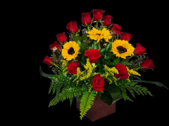 Sunlit Architecture - Bold red roses paired with sunny yellow sunflowers create a cheerful, attention-grabbing flower arrangement perfect for a birthday bouquet, a congratulations gift, or a "just because" surprise. The strong red-and-yellow contrast reads classic yet modern, so the person you're thinking of will feel celebrated and lifted the moment this arrangement arrives.  Tightly clustered roses and full sunflowers form a rounded, full display set in a clean square vase, with fern fronds and lush greenery at the base for added texture and volume. The compact silhouette makes it easy to place on a table or desk; stems are arranged for stability in the vase. For a bright everyday gift that reads both festive and heartfelt, this arrangement delivers lasting visual impact and easy care-refresh water regularly and trim stems if desired to extend bloom life.