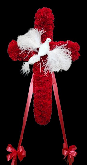 Compassion cross - A large devotional sympathy arrangement shaped as a cross, this memorial tribute is built from densely packed red carnations that create a bold, steady silhouette meant to honor a life. The cross design and ceremonial feel make it especially appropriate for a funeral service or graveside memorial, offering a clear expression of love and respect for the person you're remembering.  White feather accents and clusters of soft white carnations radiate from the center, where a white dove figurine rests-an emblem of peace and comfort. Long pink ribbon tails finish with pink bows at the base, adding a gentle, compassionate touch and a subtle contrast to the deep red. A decorative cross hard good is integrated into the design for added symbolism; overall the piece reads formal, reverent, and thoughtfully composed.  This arrangement is intended to convey sympathy and solace to family and friends at a funeral, memorial service, or graveside gathering-she'll feel honored and remembered by its dignified presence. Please check sizing and delivery options at checkout to ensure it arrives in time for services; for care, keep flowers cool and sheltered from direct sun until placement.