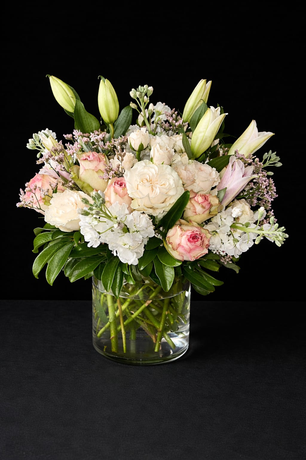 Pure Radiance - Soft cream and blush roses mingle with fragrant white lilies in this romantic bouquet-perfect as an anniversary bouquet, wedding flowers, or a gentle sympathy arrangement. The mix of roses, lilies, and white stock creates a timeless, elegant look that makes the person you're thinking of feel truly cherished and comforted.  Clusters of white stock add lush texture while unopened lily buds lift the design, giving it graceful height and a formal silhouette. Presented in a clear glass vase, the arrangement reads clean and classic; colors include white, pink, and green with a full, rounded composition that fills a room with subtle scent and refined beauty.  Whether you're marking a milestone, offering sympathy, or celebrating a wedding, this arrangement sends a message of warmth and calm. The vase holds the stems securely and tucks neatly into table settings or a mantel-no extra container needed when you're ready to gift.
