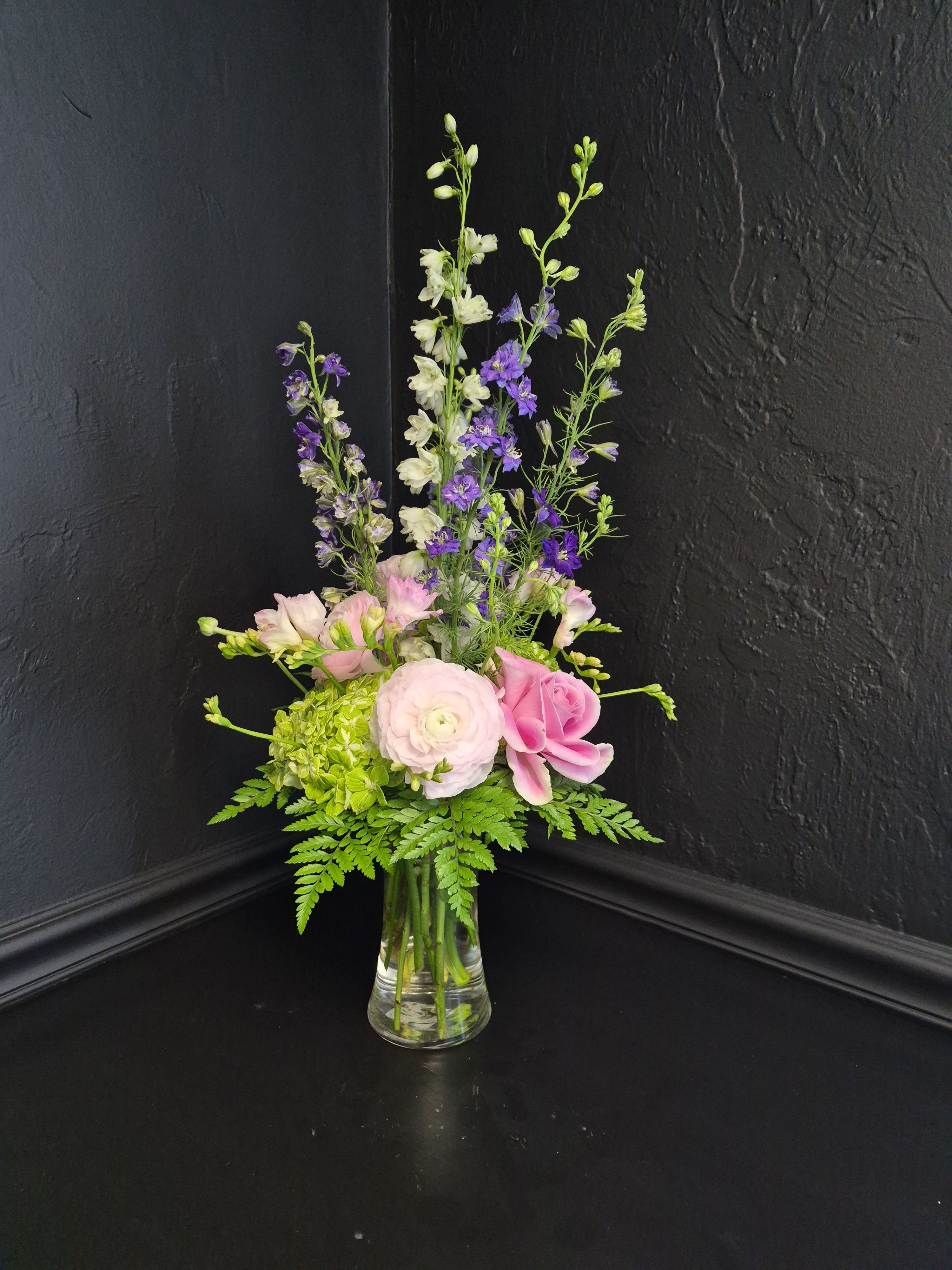 French Countryside - A tall, airy bouquet that feels like a sunlit walk through a French garden-perfect as a birthday bouquet, a spring celebration gift, or a just-because surprise. The arrangement's soft pastel palette of pink, white, purple, and green centers the recipient in a fresh, romantic moment: she'll feel seen and quietly celebrated by the gentle mix of blooms and natural movement.  Blush ranunculus and soft pink roses form the rounded, textural heart of the design while white and purple delphinium rise in slender spikes to add height and graceful movement. White freesia, green hydrangea, and airy garden foliage fill the loose, layered composition, all arranged in a clear glass vase that lets the stems and greenery show through for a simple, elegant finish. The look feels casual yet refined — ideal for thank-you gestures, springtime gifting, or brightening someone’s day.