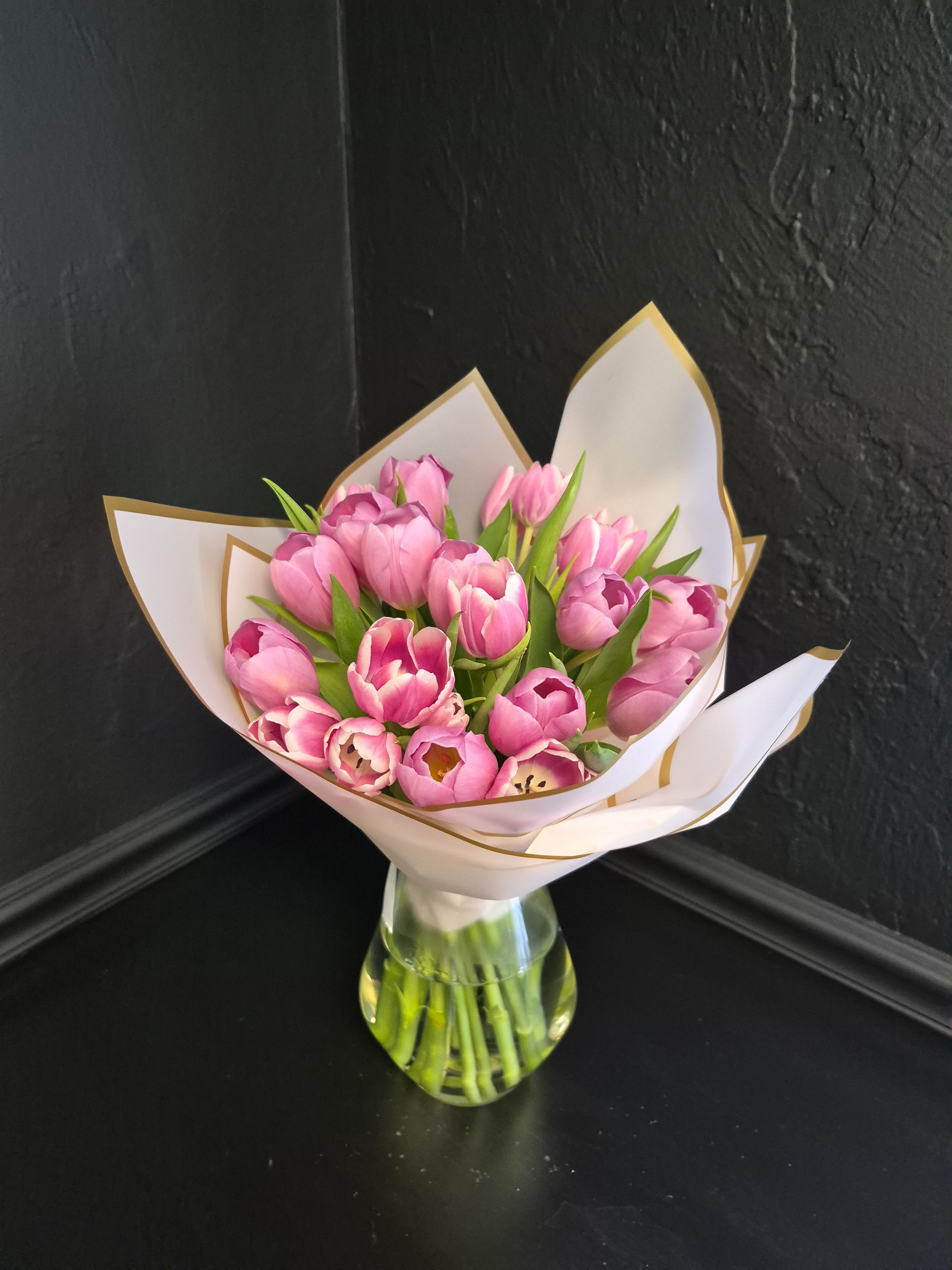 Pink Kiss - A cluster of bright pink tulips arranged in a compact, hand-tied bouquet feels like a kiss of spring - perfect as a birthday bouquet, a simple romantic bouquet, or a fresh just-because gift. The mix of open and partially closed blooms gives movement and texture so the person you're thinking of will feel both surprised and cherished the moment they see it.  Beautifully wrapped and presented in a clear glass vase, this arrangement balances vibrant pink tulips with fresh green stems for a cheerful yet elegant look. Gathered tightly for a lush, full presentation, the bouquet feels modern, romantic, and perfect for birthdays, celebrations, or thoughtful everyday gifting.  Ideal for everyday gifting, thank-you gestures, anniversaries, or spring celebrations; each bouquet arrives hand-tied and ready to display in a glass vase. For best results, recut stems and change the water every two days to keep the tulips looking their freshest.