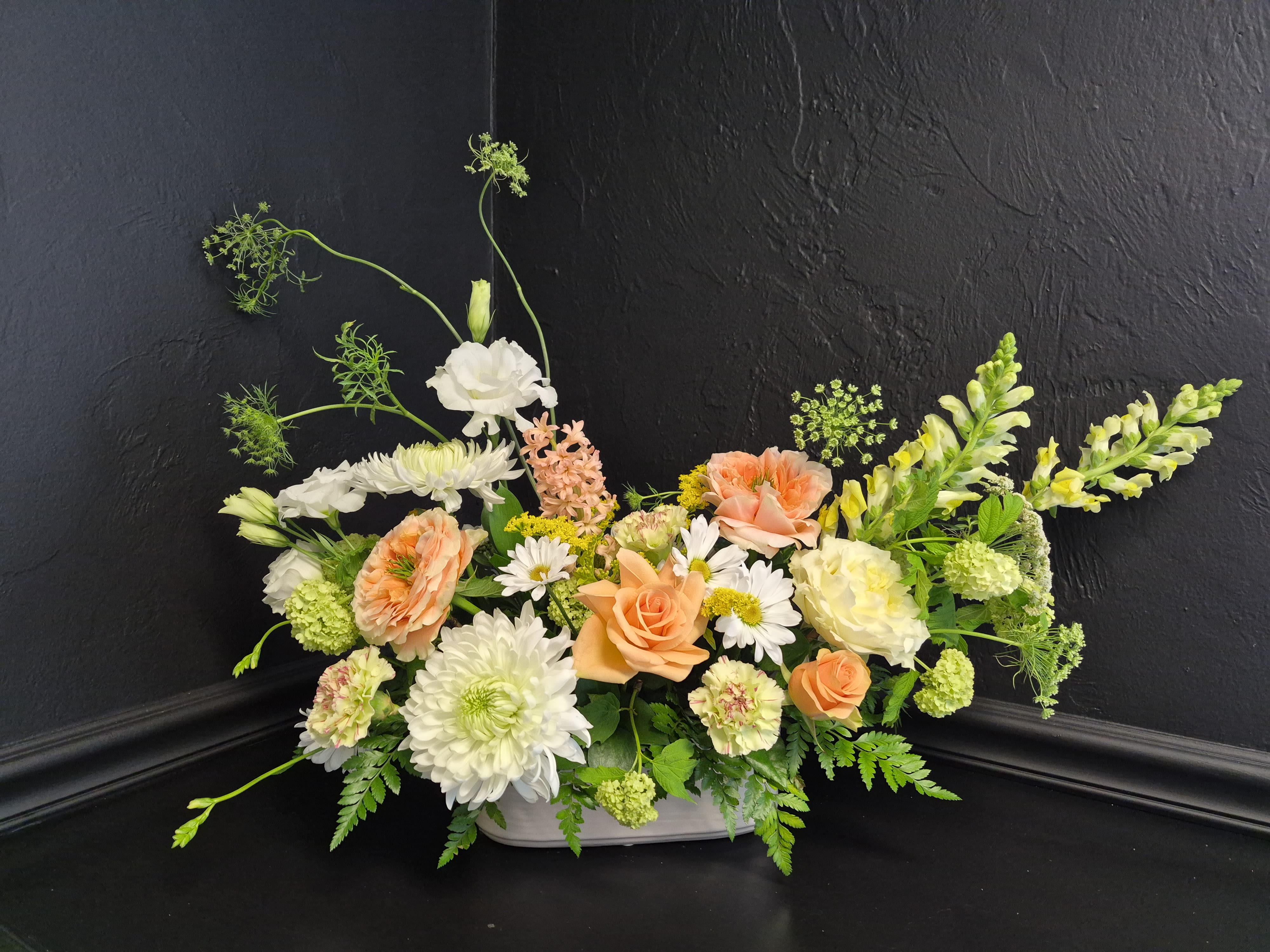 Garden Reverie - Peach garden roses and airy snapdragons spill from a low white ceramic container in a loose, garden-style arrangement — perfect for birthdays, spring celebrations, or thoughtful thank-you gifting. The soft peach, white, and fresh green palette feels elegant and uplifting, while the naturally layered silhouette creates a relaxed, effortlessly beautiful look.  This low, wide design combines lush peach garden roses with white chrysanthemums, delicate daisy-like blooms, carnations, and pale green accents for texture and depth. Long branching stems rise above the main body of the arrangement, adding graceful movement and an organic garden-inspired feel. Designed in a smooth white ceramic vessel, the bouquet also features soft yellow and blush tones that brighten and soften the overall composition.  Ideal for everyday gifting or casual celebrations, this arrangement brings a fresh, garden-inspired atmosphere to kitchen tables, desks, or bedside spaces. Its compact yet airy shape makes it easy to display, while the layered seasonal blooms create a full, abundant presentation. To keep the arrangement fresh, simply add a small amount of water to the floral foam every couple of days as needed.