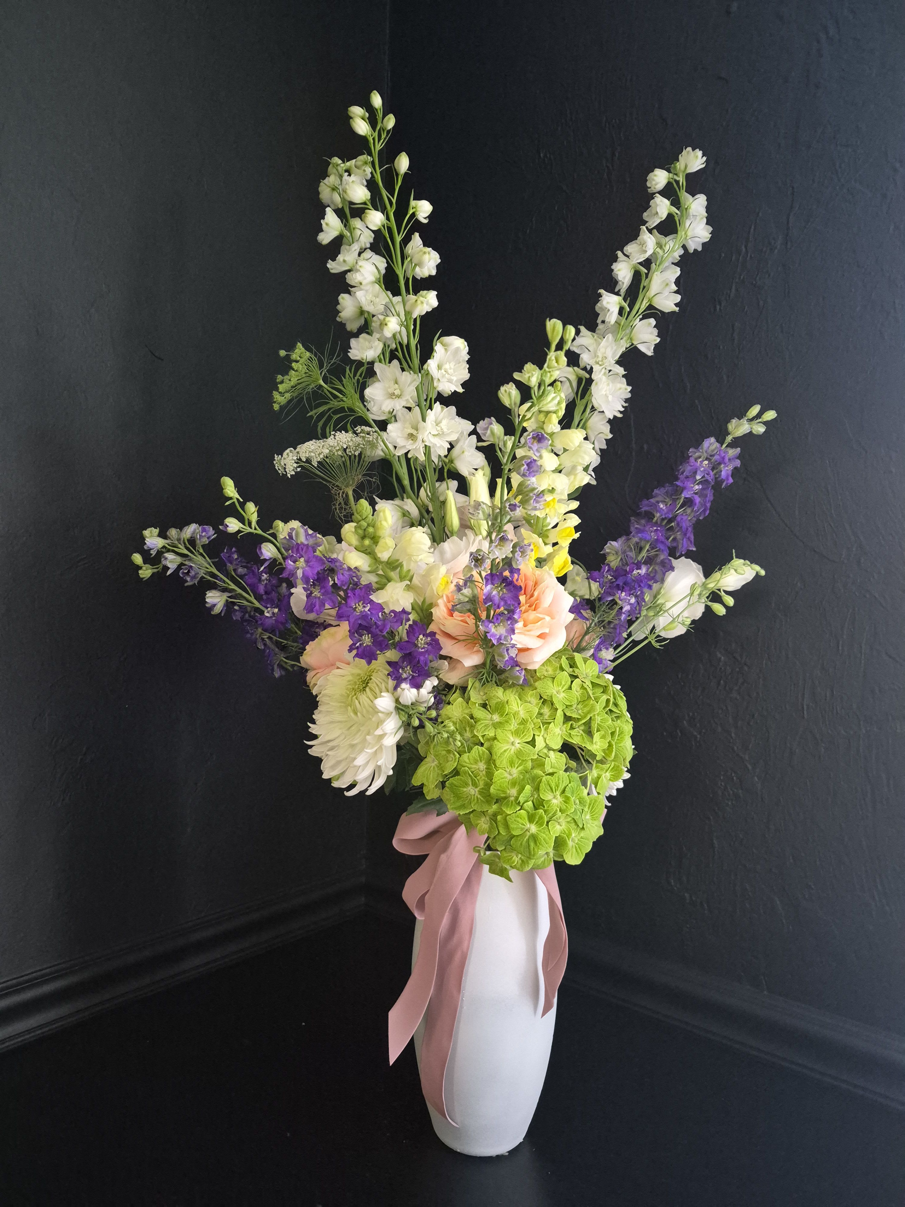 Spring Elegance - Tall purple delphinium spires rise above a lush base of green hydrangeas and soft pink and white blooms, creating an airy spring arrangement perfect for birthdays, thank-you gifts, or elegant seasonal celebrations. Its graceful vertical design gives the piece a refined, romantic character that makes the recipient feel thoughtfully celebrated without overwhelming the space.  This arrangement features vibrant purple delphinium paired with fresh green hydrangea clusters, pale pink carnations, white chrysanthemums, and delicate seasonal filler blooms, all gathered in a sleek white ceramic vase finished with a soft pink ribbon. The tall silhouette and layered textures create a balanced composition with movement, height, and a garden-inspired elegance.  Perfect for anniversaries, hostess gifts, spring gatherings, or thoughtful “thinking of you” moments, this arrangement brings a fresh and sophisticated presence to entryways, dining tables, or office spaces. To keep blooms looking their best, refresh the water regularly, trim stems every few days, and place away from direct heat or sunlight.