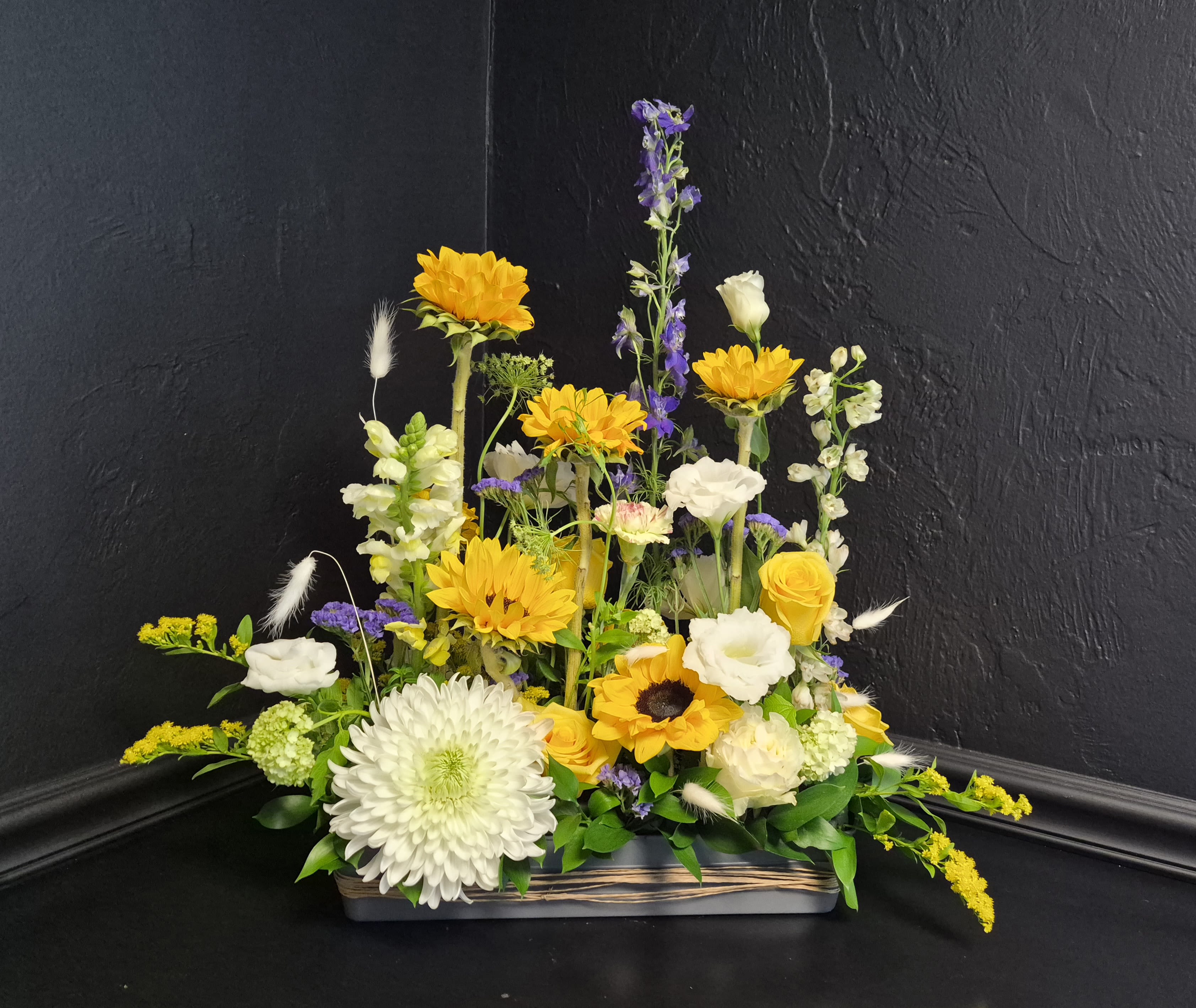 Golden Ascent - Sunflowers rise above a lush mix of white chrysanthemum, creamy lisianthus, yellow roses, purple and white delphinium, snapdragons, and fresh seasonal greenery in this bright garden-style arrangement. Designed low and wide in a modern rectangular container, "Golden Ascent" captures the feeling of blooms gently reaching upward toward the light, with airy movement, layered textures, and soft decorative grasses creating a naturally abundant look.  With its sunny yellow palette, crisp whites, and touches of soft violet, this arrangement feels cheerful, uplifting, and elegant at the same time — perfect for birthdays, celebrations, thank-you gifts, or simply bringing warmth and light into someone's space. Ready to display and designed to brighten any table or entryway with fresh garden-inspired charm.