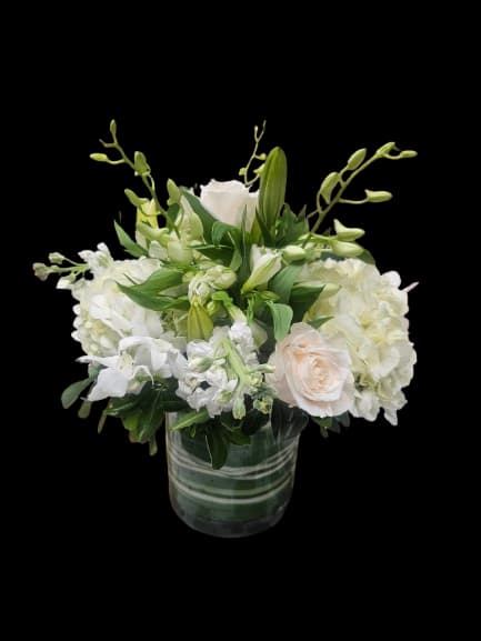 Heavenly - Soft white blooms create a calm, elegant centerpiece perfect for birthdays, thank-you gifts, sympathy gestures, or refined everyday occasions. Rounded white hydrangeas and creamy white roses form the heart of the arrangement, while white stock, alstroemeria, lily buds, and delicate dendrobium orchids add softness, texture, and graceful movement for a design that feels peaceful, fresh, and quietly luxurious.  Arranged in a clear glass vase, the bouquet balances lush hydrangea clusters, velvety roses, sculptural orchid sprays, fragrant stock, and lilies that gradually open over time for an arrangement that continues to evolve beautifully over several days. The all-white palette with touches of soft green creates a timeless, versatile look that suits elegant homes, offices, dining tables, or bedside spaces.  Designed to bring a sense of serenity and beauty, this arrangement is ideal for anyone who appreciates classic floral styling with a fresh garden-inspired feel. Vase included; refresh water regularly and trim stems slightly every few days to prolong bloom life.