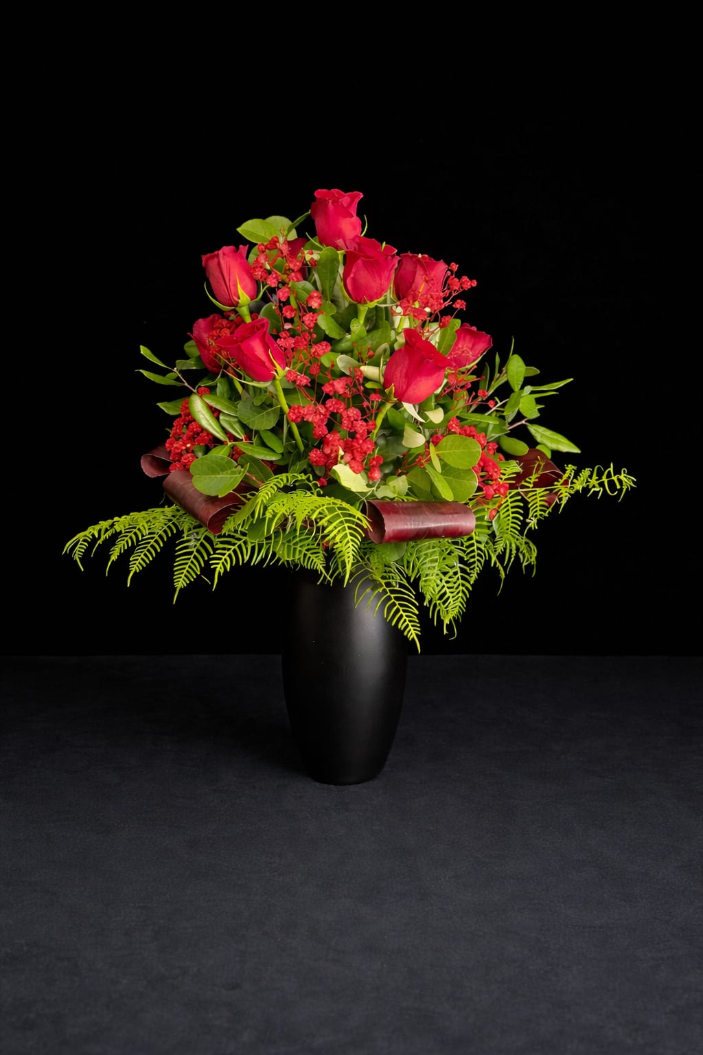 Red Velvet Dream - Vivid red roses gathered in a tall black vase create an immediate, romantic impact-an ideal birthday bouquet, anniversary arrangement, or thoughtful thank-you gift that will make the person you're thinking of feel cherished. The combination of bright red blooms and burgundy ribbon gives a polished, elegant look that reads both celebratory and intimate.  A classic dozen roses form the heart of this arrangement, accented with small red filler flowers and airy, fern-like greenery that spills outward for a layered, lush silhouette. The roses sit in a sleek black vase that heightens the color contrast and keeps the presentation modern and refined; burgundy ribbon loops add a warm, textural finishing touch.  Perfect for gifting across many occasions, this arrangement suits someone who loves romantic, feminine color with contemporary styling. Vase-ready for immediate display-refresh the water every few days and trim stems if desired to extend bloom life.