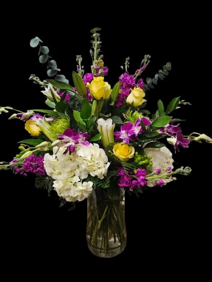 Opera - A bold cascade of yellow roses, white hydrangeas, purple Dendrobium orchids, and elegant white lilies creates an elevated, celebratory centerpiece — perfect as a birthday bouquet, anniversary arrangement, or thoughtful thank-you gift. The Spring Opera flower arrangement features a lively yet balanced palette of bright yellow, crisp white, and rich magenta-purple tones designed to make someone feel truly celebrated.  Arranged in a clear glass vase, this design is lush and layered with airy stems, branching accents, and deep green foliage that create a dramatic, upscale silhouette with graceful movement. The composition combines voluminous hydrangea blooms, classic roses, exotic orchids, and sculptural lilies for rich texture, dimension, and height. Sophisticated yet vibrant, this arrangement brings warmth, elegance, and modern garden-inspired style to any home or office.  Care: Refresh water regularly and trim stems every few days to extend bloom life.