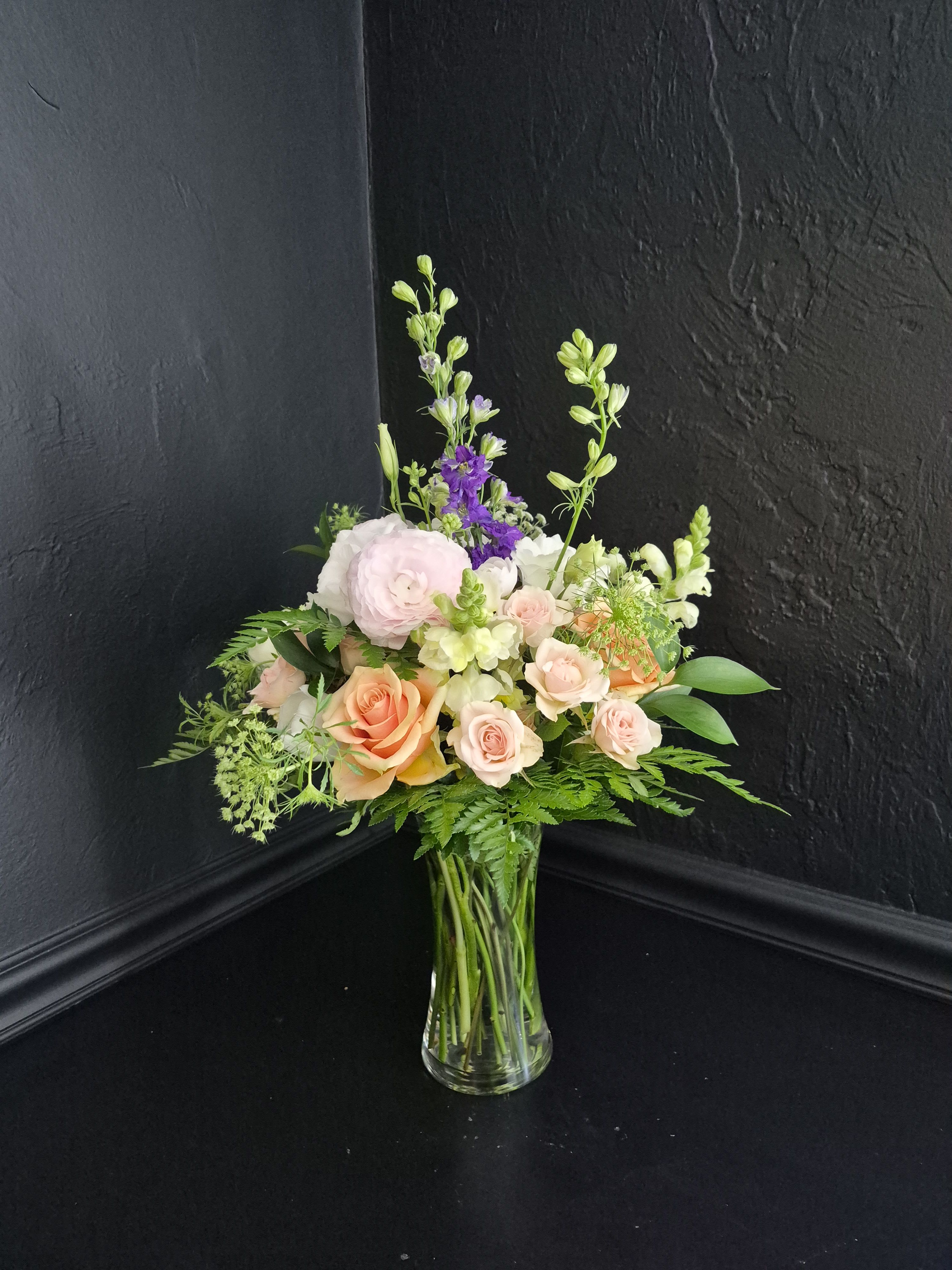 Spring Poetry - Soft blush roses and tall lavender delphinium create a gentle, garden-fresh statement in this romantic spring arrangement — perfect for birthdays, anniversaries, thank-you gifts, or thoughtful everyday moments. The blend of blush pink, peach, white, and soft purple tones feels fresh, airy, and uplifting, creating a design that makes the recipient feel quietly celebrated and cared for.  Roses form the rounded heart of the arrangement while delicate delphinium and snapdragon stems add height and movement to the airy, layered silhouette. Ranunculus, lisianthus, and white tulips bring softness and texture to the arrangement, while fern-like greenery and airy filler flowers add movement and a natural garden-inspired feel. All blooms are arranged in a clear glass vase for an elegant yet effortless presentation that fits beautifully on dining tables, entryways, bedside tables, or office spaces.  Perfect for spring gifting or simply brightening someone’s day, this arrangement balances romantic softness with natural elegance. To keep blooms looking fresh, change the water every two days, trim stems slightly at an angle, and keep the arrangement away from direct sunlight and heat.