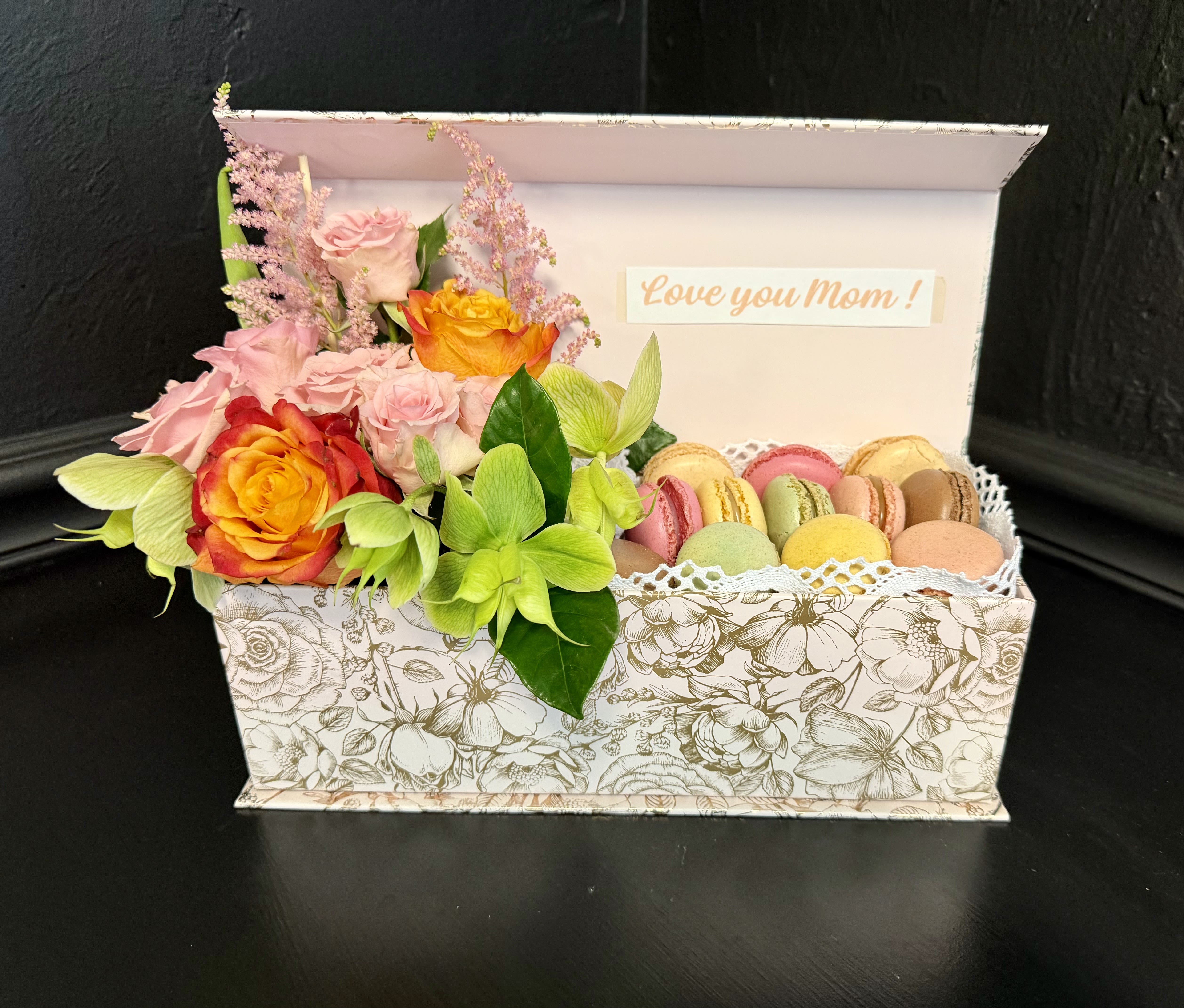 Sweet Mom - Bright clusters of orange roses paired with orange alstroemeria and yellow tulips bring a sunny, affectionate energy to this Mother's Day gift set - a cheerful gift box and macarons combo perfect for a birthday or to say "I'm thinking of you." The compact presentation feels celebratory and personal, with a small printed card that reads "Love you Mom!" to make the recipient feel truly seen and loved.  The arrangement sits on one side of a patterned white decorative gift box, the blooms accented by pink and white highlights against a soft paper liner and finished with a ribbon label. On the other side, a tray of pastel macarons adds a delicate texture and taste contrast to the vivid floral colors; the mixed-color palette of pink, orange, yellow and white makes the set bright and versatile for spring or any joyful occasion.  Designed as a ready-to-gift keepsake, this set is ideal for moms, grandmothers, or anyone who deserves a sunny surprise - it reads as both thoughtful and festive. The compact box is presentation-ready; keep flowers in a cool spot away from direct sunlight and enjoy the macarons fresh from the tray for best flavor.