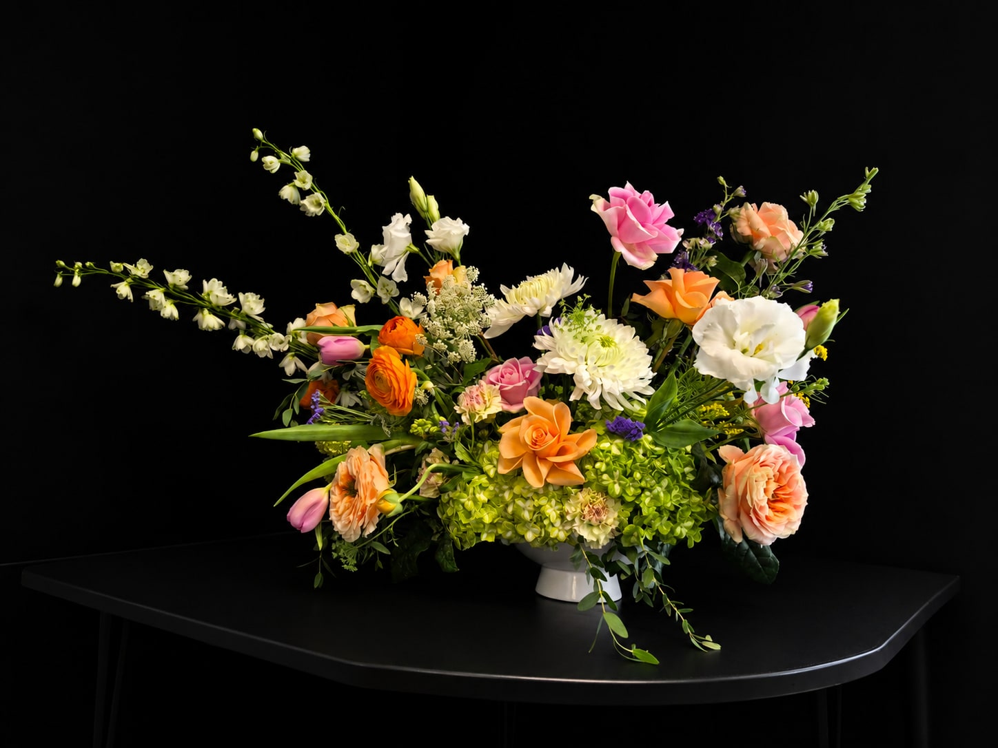 Mom's Favorite - Bright, airy, and joyfully gathered — this garden-style flower arrangement is designed to make Mom feel truly celebrated. With its soft pastel blooms, cheerful citrus tones, and naturally flowing shape, it’s perfect as a birthday bouquet, Mother’s Day gift, or a heartfelt “just because” surprise. Romantic yet fresh, it brings together elegant garden flowers and lush seasonal textures for a look that feels both uplifting and comforting.  A lush mix of roses, lisianthus, ranunculus, chrysanthemums, tulips, carnations, delphinium, hydrangea , snowball viburnum and other seasonal blooms creates layered texture and movement. Saturated oranges and pinks rise in spiky vertical accents above a broad base of glossy green foliage, while peach, white and lime-green blooms plus small purple and yellow fillers add playful contrast. The result reads both bold and airy-rounded focal flowers set against long branching stems for a lively, hand-arranged look.  Arranged in a white pedestal bowl for an elegant tabletop presence, this design suits celebrations, thank-you gifts, birthdays and spring moments alike-she'll feel celebrated the minute she sees it. Designed to be a full, visible display, it arrives ready to enjoy; refresh the water every other day and trim stems as needed to keep blooms vibrant.