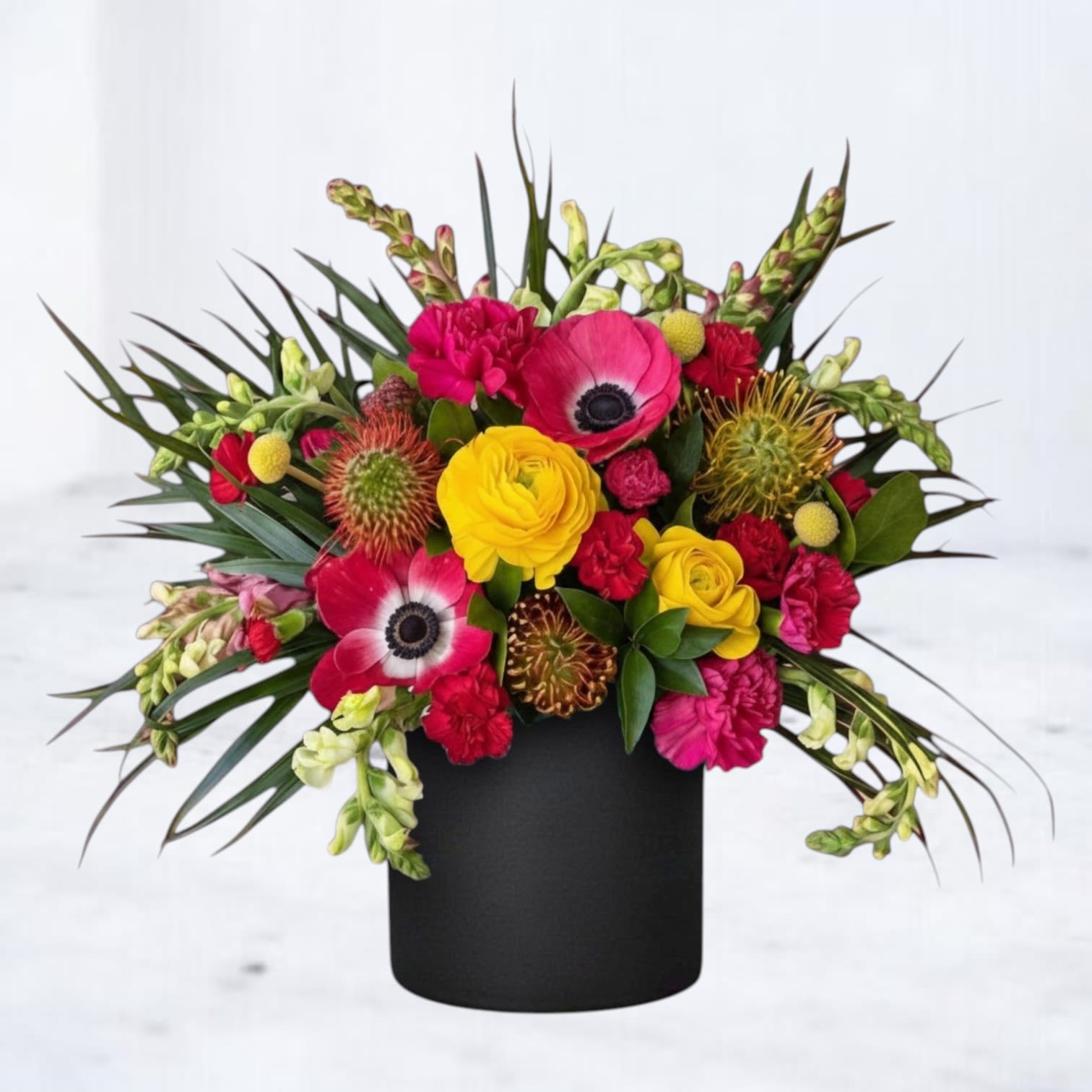 Calyx - Calyx is a bold, modern floral arrangement designed with striking color contrast, sculptural texture, and dynamic movement. Featuring vibrant ranunculus, anemones, specialty blooms, and unique textural elements, this arrangement is artfully composed with structured greenery to create a dramatic, design-forward look.  Designed in a matte black vessel, Calyx blends saturated reds, hot pinks, and vivid yellows with deep green foliage for a composition that feels edgy, contemporary, and visually captivating. The combination of bold florals and spiky textures gives this arrangement a sculptural presence, making it a true statement piece.  Perfect for birthdays, celebrations, modern interiors, or sending something unexpected and unforgettable, this vibrant, high-impact flower arrangement is ideal for those who love unique, artistic floral design.  Same-day flower delivery is available in Magnolia, TX, The Woodlands, Montgomery, Conroe, Spring, Tomball, and surrounding North Houston areas.  Due to seasonal availability, some blooms may be replaced with similar varieties of equal or greater value to maintain the design’s look and integrity. Your arrangement will always reflect the same color palette, style, and premium quality.