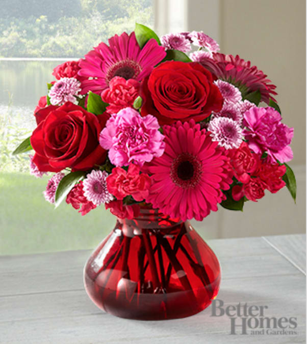 The FTD® Make Me Blush Bouquet by Better Homes and Gardens® - VASE INCLUDED - Picked fresh from the farm to make this February 14th a day full of surprises and romance, the Better Homes and Gardens® Make Me Blush Valentine's Day Bouquet is boldly sweet with blooming beauty. Hand gathered at select floral farms and bringing together an array of blooms in varying shades of pink and red, this stunning mixed flower bouquet has been picked fresh for you to make your special someone feel the love on this truly romantic day. Bouquet includes: hot pink gerbera daisies, red roses, hot pink carnations, hot pink mini carnations, lavender button poms, and lush greens. Presented in a modern red glass posie vase.  GOOD bouquet is approx. 11"H x 11"W.  BETTER bouquet is approx. 12"H x 12"W. BEST bouquet is approx. 13"H x 14"W. Your purchase includes a complimentary personalized gift message.