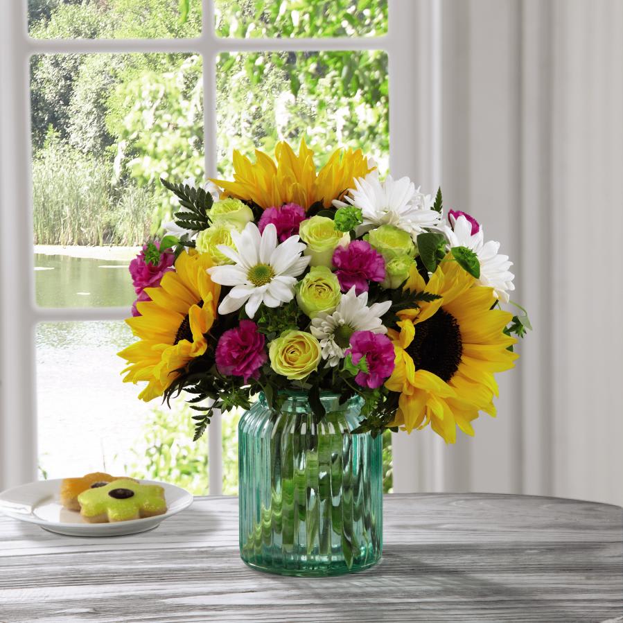 The FTD Sunlit Meadows Bouquet by Better Homes and Gardens - Deluxe - FTDÂ¨ proudly presents the Better Homes and GardensÂ¨ Sunlit MeadowsÂª Bouquet. Blossoming with every color you are drawn to during spring's arrival, this gorgeous bouquet plays with cool and warm tones to draw you in and create that perfect way to touch your recipient's heart. Gorgeous sunflowers are the standout bloom in this arrangement offset by yellow spray roses, magenta mini carnations, white daisies, and bupleurum, all perfectly situated in a keepsake aqua glass vase to compliment and contrast with the flowers sunlit beauty. A wonderful Mother's Day, Easter, or happy birthday gift! GOOD bouquet includes 11 stems. Approx. 12"H x 12"W. BETTER bouquet includes 13 stems. Approx. 12"H x 13"W. BEST bouquet includes 18 stems. Approx. 13"H x 15"W. EXQUISITE bouquet includes 21 stems. Approx. 13"H x 15"W.