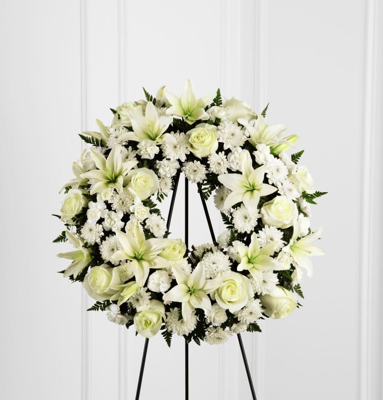 FTD Treasure Tribute Wreath - The FTD Treasured Tribute Wreath offers peaceful wishes of   heartfelt sympathy with each delicate bloom. Bright white roses, Asiatic   lilies, mini carnations and cushion poms are beautifully arranged to   form an elegant accented with lush and vibrant greens. Displayed on a   wire easel, this gorgeous tribute is a wonderful symbol of eternal life   and sweet serenity. Approximately 22 inches in diameter.  no dimensions