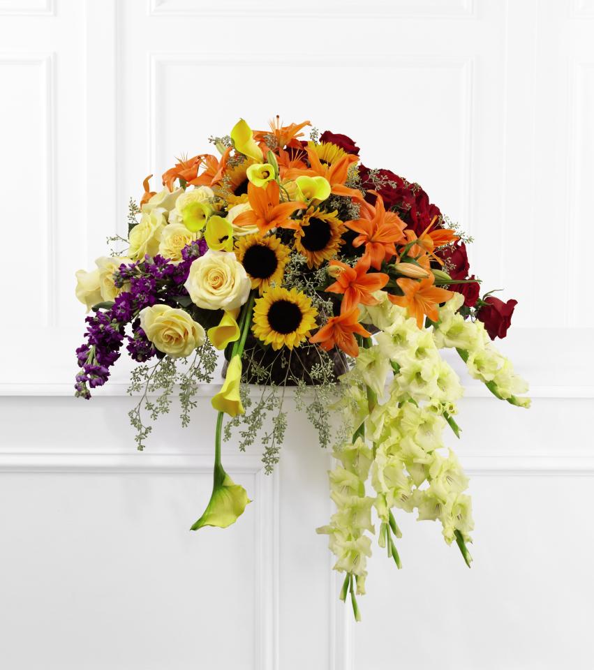 FTD Peaceful Tribute Arrangement - The FTD Peaceful Tribute Arrangement is an elegant memorial that   bursts with sun-kissed beauty. This three-sided arrangement brings   together the varied elements, colors and textures of cream and red   roses, purple stock, yellow mini calla lilies, orange Asiatic lilies,   yellow gladiolus, sunflowers and lush greens to create a bright and   brilliant display. Seated in a large round banana leaf basket, this   arrangement will convey warm sentiments at their final farewell service.      30h x 32w