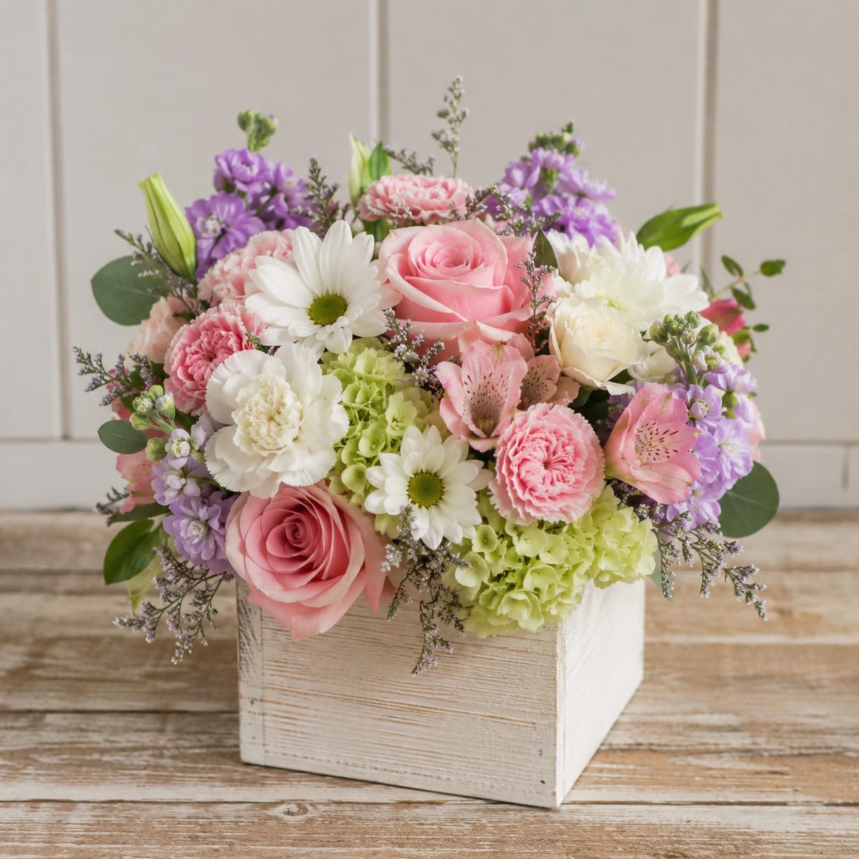 Paste Box of Blooms by Petal Perfection Flower Company  - Soft pink roses and pale green hydrangea form the heart of this pastel birthday bouquet, arranged in a white wooden box that feels like a little garden on the table. The rounded, abundant shape-soft pinks, clean whites, and touches of purple-creates a gentle, romantic look ideal for anniversaries, spring celebrations, or a thoughtful "thinking of you" gift for someone who loves soft color palettes.  This compact arrangement pairs roses, carnations, daisies, hydrangeas, alstroemeria, and a few lily buds for height and a fresh, layered texture. The flowers are densely layered for a full, plush appearance, while airy filler stems give a lightly loose, natural finish. The sturdy white wooden box makes it easy to display on a desk or mantel; order now to send a pastel-sweet bouquet that will make the recipient feel truly seen.