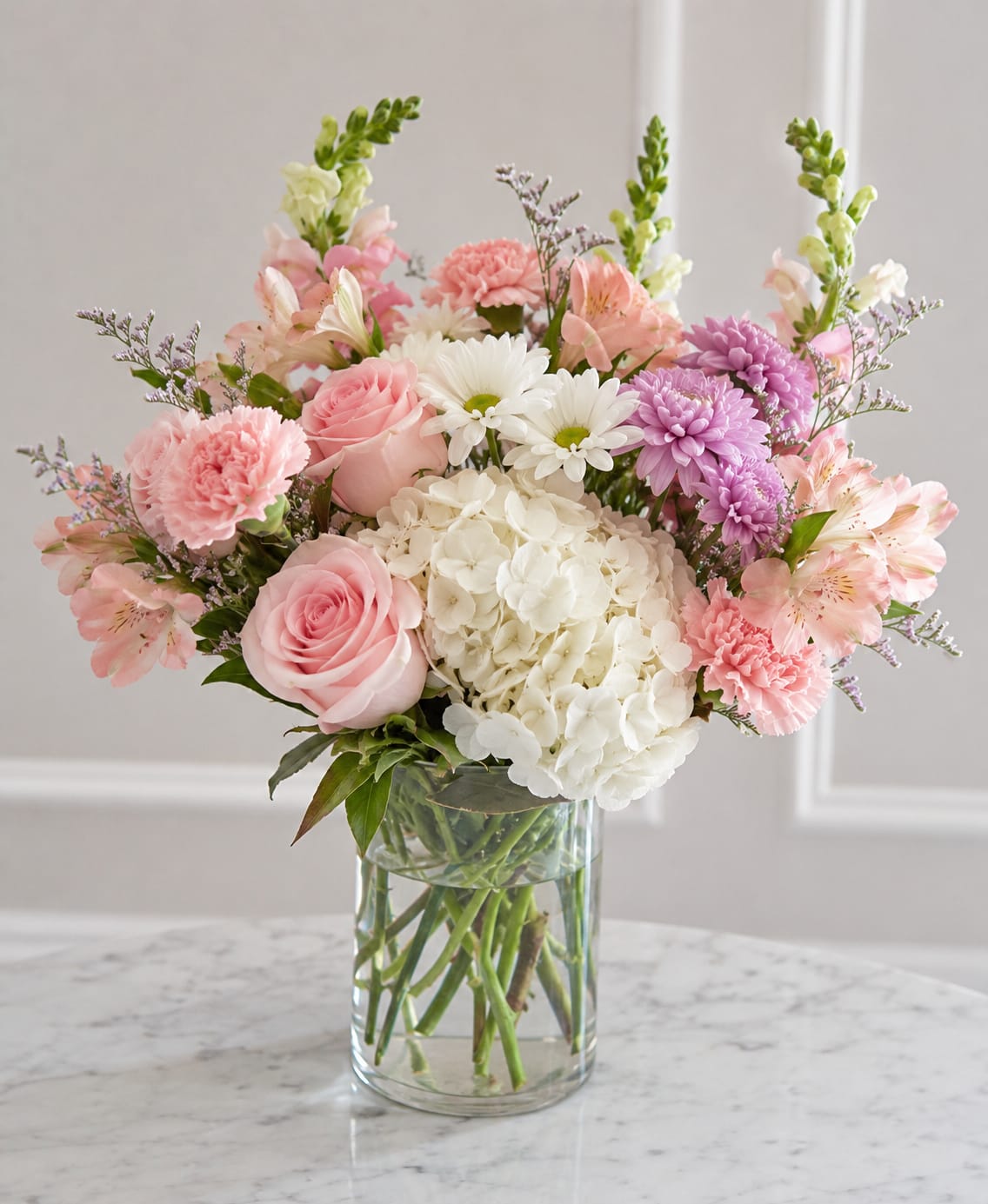 Petal Perfection's Perfectly Pink And White - A soft, romantic spray of pink and white blooms makes an immediate, joyful impression - perfect for a birthday bouquet, Mother's Day gift, or a tender anniversary arrangement. The rounded composition centers on a lush white hydrangea cluster that gives the design an airy, garden-fresh feel; surrounding pink roses and carnations read as affectionate and sincere while lavender-purple mums and white daisy-like blooms add delicate contrast.  This arrangement includes roses, hydrangeas, carnations, daisies, and alstroemeria in a pastel palette of pink, white, and purple, presented in a clear cylindrical glass vase so stems show through the water for a clean, classic look. The full, compact shape is easy to display on a table or bedside, and the mixed textures - velvety rose petals, ruffled hydrangea clusters, and dainty daisies - create a tactile, invitation-to-smile effect.  Choose this bouquet when you want her to feel genuinely seen and celebrated; it reads as thoughtful, feminine, and fresh for springtime gifting or anytime you want to say "you're special." Care is simple: keep water fresh, trim stems every few days, and place away from direct heat to extend bloom life.
