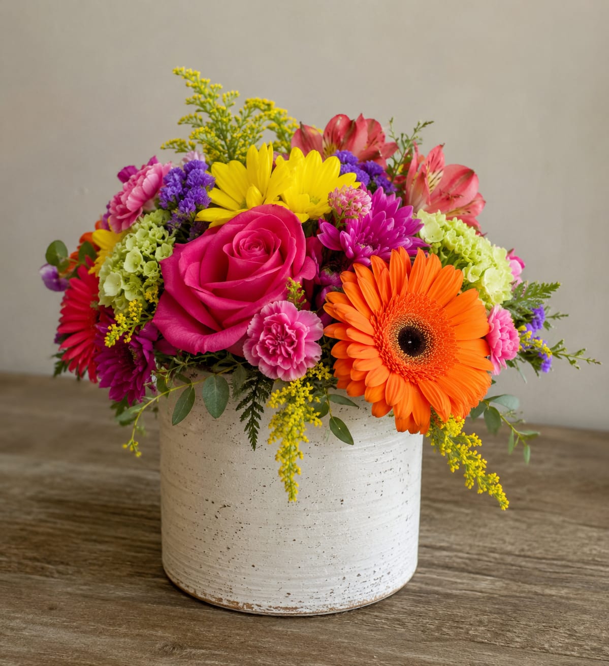 Bold and Beautiful by Petal Perfection Flower Company  - A bright pink rose and a bold orange gerbera daisy take center stage in this cheerful birthday bouquet, making Bold and Beautiful an ideal flower arrangement for birthdays, congratulations, or just to brighten someone's day. The low, rounded composition in a white ceramic pot reads like a festive tabletop centerpiece-perfect for a kitchen table, office desk, or mantle-so the person you're thinking of will feel instantly celebrated by the saturated color and joyful energy.  This dense mixed bouquet combines roses, gerbera daisies, carnations, chrysanthemums, alstroemeria and seasonal filler blooms in a palette of pink, orange, yellow, purple and lime tones. The mix of large focal flowers and varied textures-soft rose petals, graphic gerbera faces, ruffled carnations and daisy-like mums-creates a lively, hand-gathered look that stays compact in its white ceramic pot. The low-profile design is arranged to read well from all sides and makes a bright, long-lasting focal point.  Perfect for springtime parties, birthday surprises, or "thinking of you" deliveries, this arrangement is designed to make the recipient feel noticed and joyful. The ceramic pot keeps it stable for tabletop display; for best freshness, place in a cool spot away from direct heat and refresh water as needed according to standard floral care.