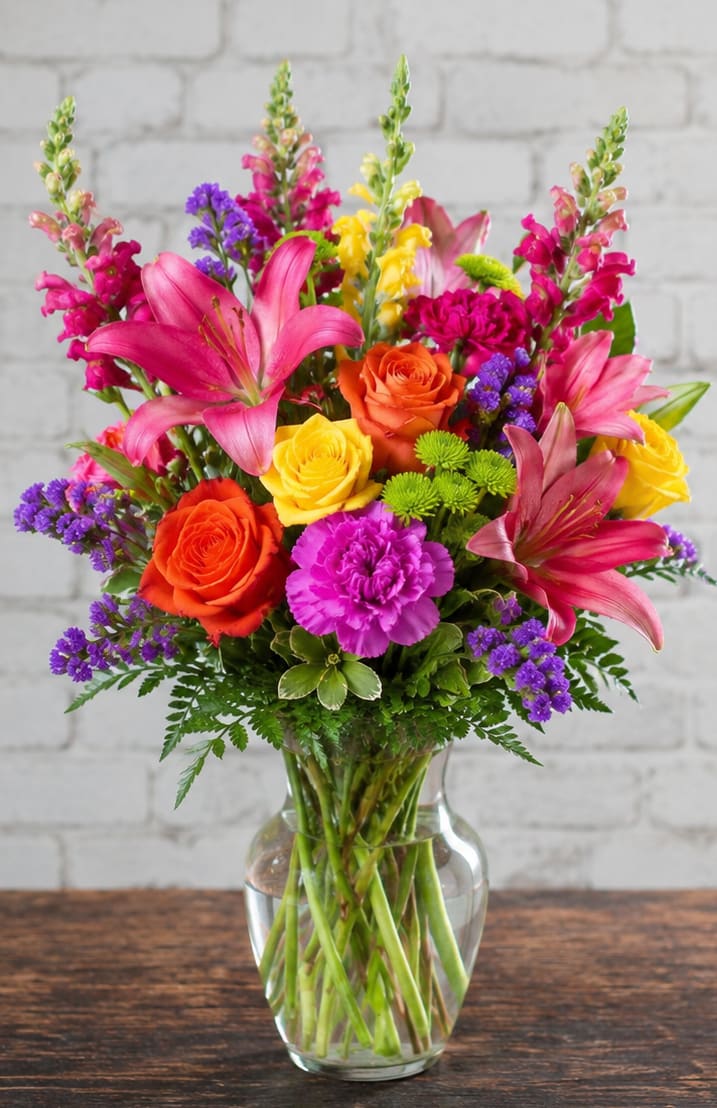 Petal Perfections Colors of the Rainbow  - Vivid pink lilies and tall snapdragon spikes create an uplifting focal point in this birthday bouquet-style flower arrangement for celebratory occasions. The Colors of the Rainbow arrangement reads like a joyful party in bloom-perfect for birthdays, get-well wishes, or simply letting someone know you're thinking of them-so the person you send it to will feel genuinely celebrated the moment they see it.  This full, upright design pairs orange and yellow roses with magenta carnations and purple filler flowers for a layered, lively silhouette. Arranged in a clear glass vase, the composition mixes rounded blooms and vertical snapdragons to add depth and movement while keeping a fresh, springlike palette. The finished bouquet arrives ready to display; refresh the water every few days, recut stems at an angle if needed, and enjoy bright color that lasts through the week.