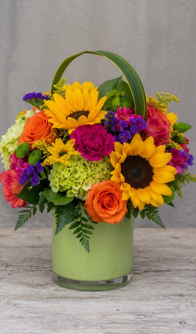 Color Pop for Apollo Beach - Two bold sunflowers take center stage in this lively flower arrangement, making it an ideal birthday bouquet, congratulations gift, or cheerful just-because surprise for someone in Apollo Beach. The mix of yellow, pink, orange, and purple creates an energetic color pop that reads as joyful and celebratory - the kind of arrangement that makes the recipient feel instantly noticed and uplifted.  Surrounding the sunflowers are vibrant orange roses, bright pink carnations, soft green hydrangea, and clusters of purple filler blooms, arranged into a full, rounded composition. The flowers sit in a light green glass vase with a curved handle that gives the piece a basket-like, casual charm while keeping the profile neat enough for tabletops or entryway consoles. Textures range from the sunflowers' bold centers to the roses' layered petals and the hydrangea's billowy clusters, delivering visual depth from every angle.  Perfect for marking birthdays, sending congratulations, or bringing a burst of sunshine to someone who needs cheering, this arrangement reads as festive and upbeat without being over the top. Order with confidence for same-week delivery where available; keep flowers in fresh water, away from direct heat, and trim stems slightly if you'd like to refresh the arrangement and extend its lively presence.