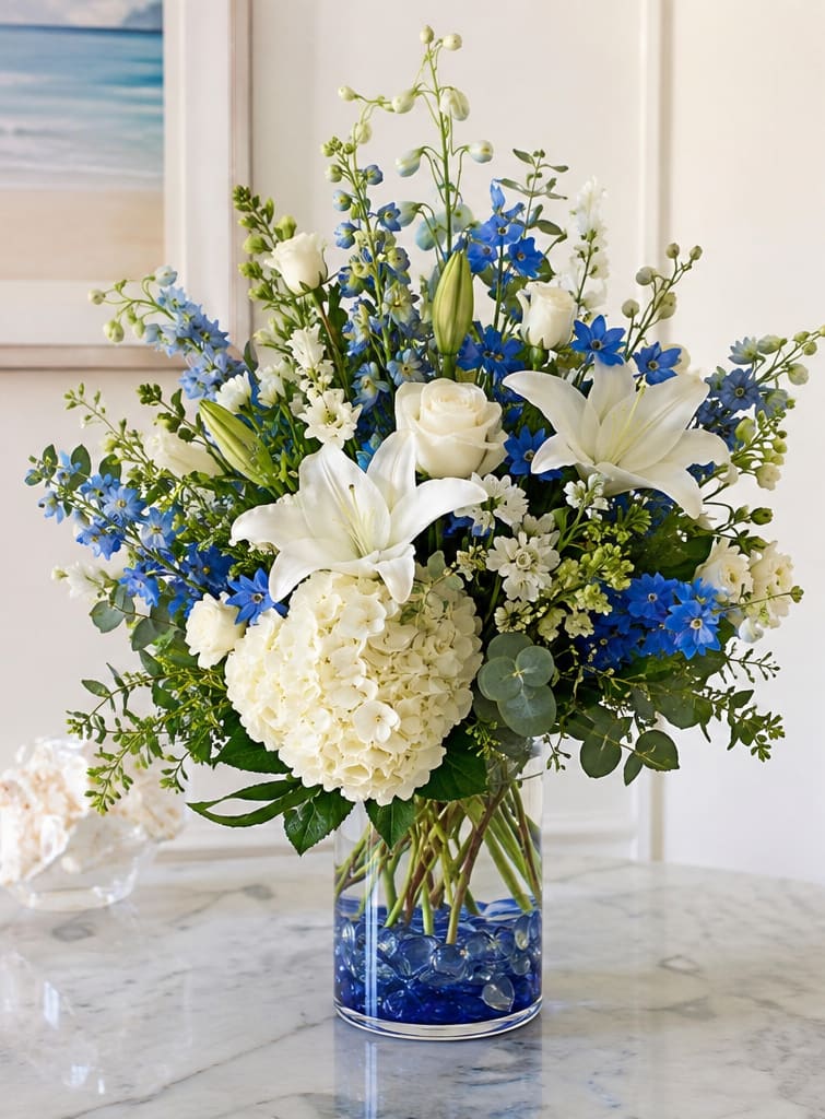 River's Edge for Riverview - Crisp white lilies and roses meet vivid blue delphinium and accent blooms in a tall, airy flower arrangement perfect for sympathy, celebration, or a refined home display. This white-and-blue flower arrangement for everyday elegance places the occasion - whether a thoughtful condolence or a graceful centerpiece for a special moment - on the person you're thinking of, helping them feel comforted and seen.  The design features large white lilies, white roses, and a rounded white hydrangea bloom supported by vivid blue accent flowers and greenery, arranged in a clear cylindrical glass vase. Stems rise above a fuller base for a vertical, elegant silhouette, and blue glass stones at the bottom add a cool, river-like shimmer that enhances the white-and-blue contrast.  Ideal for sending to someone who appreciates refined, fresh floral styling, this arrangement works as a sympathy tribute, a celebration piece, or an understated statement for a living room or entry. Delivered in a clear glass vase ready to display - standard care is to refresh the water every few days and trim stems as needed to keep the lilies and roses looking their best.