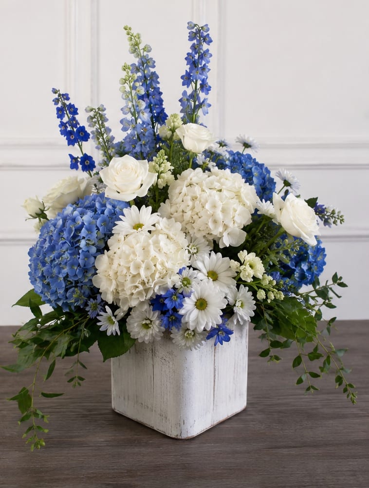 Wimauma Blue No More - A tall, layered sympathy arrangement that reads as both formal and softly comforting, this white and blue flower arrangement is ideal for a funeral, wedding, or elegant everyday display. The upright composition features large white hydrangea blooms alongside vivid blue hydrangeas and tall blue delphinium spikes, creating a cool, airy palette that naturally conveys calm and grace to the person you're honoring.  White roses and daisy-like blossoms soften the stronger blue accents, adding gentle texture and romantic detail amid the bold hydrangea heads. The flowers are arranged in a distressed white wooden container that lends a subtle rustic finish while keeping the overall look refined and structured. Sized to stand out without overwhelming a space, it's the kind of arrangement that makes someone feel seen and comforted; place in a cool area away from direct sunlight and refresh water as needed to prolong freshness.
