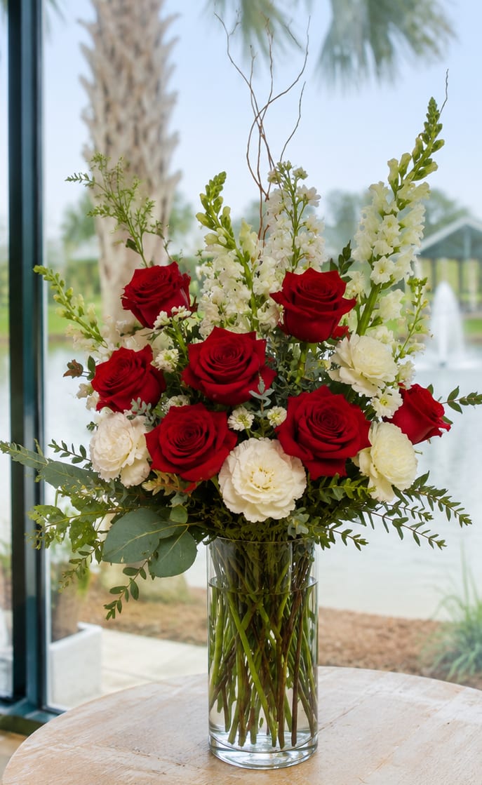 Forever In My Heart  Ellenton  - A striking upright display of vivid red roses and creamy white blooms designed to convey deep affection and timeless remembrance-perfect as an anniversary bouquet, wedding centerpiece, or sympathy arrangement. The bold red-and-white contrast creates an elegant, formal impression that will make the person you're thinking of feel truly honored and seen.  This flower arrangement pairs long-stem red roses with white carnations and other creamy white flowers, arranged densely at the center for a lush, full look. Airy white spikes and branching greenery extend upward to add height and graceful movement, all presented in a clear cylindrical glass vase that highlights the clean lines and classic color palette. The composition reads romantic yet refined, with rich textures from velvety roses and soft carnation petals.  Choose this for moments that call for heartfelt emotion-celebrating lasting love, marking a solemn remembrance, or elevating a wedding table. The vase sizing and sturdy stems make it easy to display; change the water every other day and trim stems slightly for best longevity. Order with confidence to send a message that feels both elegant and deeply personal.