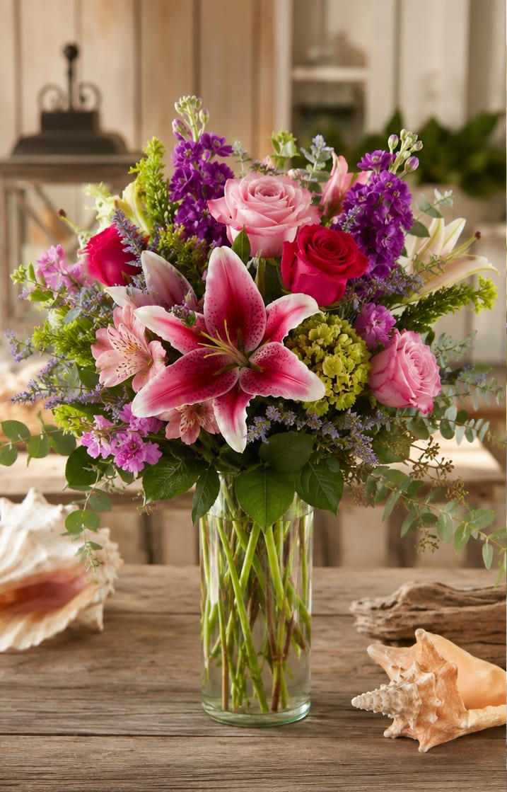 Sea Side Romance for Tampa Bay - A lush garden-style arrangement that feels like a coastal breeze-this flower arrangement for birthdays, anniversaries, Mother's Day, or any celebratory moment pairs pink roses with a prominent pink-and-white lily to create a romantic focal point that will make the person you're thinking of feel cherished. The lively mix of bright pinks and purples lends an immediate sense of joy and warmth, while touches of white and green keep the look fresh and elegant.  Handcrafted in a clear cylindrical glass vase, the design layers roses, lilies, alstroemeria, stock, and hydrangeas into a full, rounded composition with clusters of purple and blush filler blooms for texture and depth. The result is dense and lush without feeling fussy-perfect for someone who loves rich color and a garden-inspired silhouette.  Ideal as a heartfelt birthday surprise, anniversary tribute, or Mother's Day gift, this bouquet sends a message of affection and celebration that reads as both romantic and upbeat. Delivered ready to enjoy in its glass vase, it brightens a dining table, entryway, or bedside; simple care-fresh water and trimming stems every few days-keeps blooms looking their best.