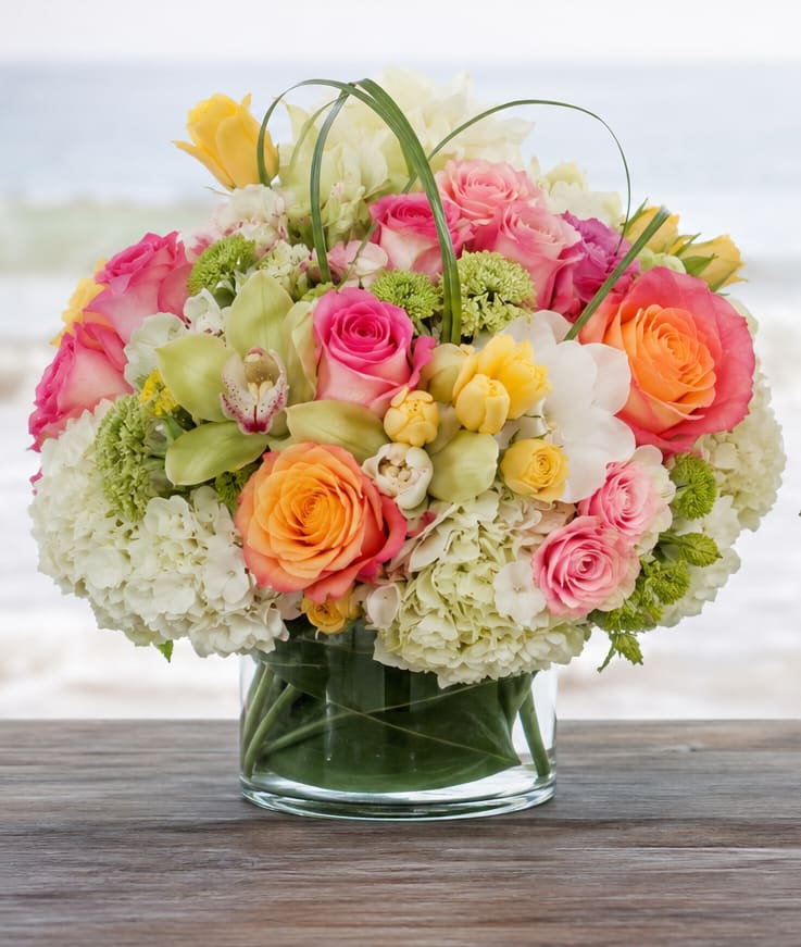 Bayshore Elegance  - Soft pastel abundance greets the recipient at first glance - a cheerful birthday bouquet or Mother's Day arrangement that reads both elegant and joyful. This flower arrangement for birthdays, anniversaries, or spring celebrations centers on a rounded, full composition that makes the person you're thinking of feel truly celebrated and seen.  Large clusters of pink and white hydrangeas create a lush, cloud-like base while peach and yellow roses weave warmth and classic romance through the design. Delicate orchid blooms add a silky, unexpected texture and height, all presented in a simple clear glass vase that showcases the stems and keeps the focus on the blooms. The palette of pink, white, yellow, and orange offers a bright, pastel look that complements warm-weather occasions and brings a fresh, uplifting presence to any room.  Perfect for sending to a mom, partner, or friend to mark an anniversary or to brighten a springtime table, this arrangement arrives full and abundant to make an immediate impression. The clear glass vase is ready to display; fresh-cut stems benefit from daily water checks and a fresh trim every few days to extend vase life and keep the display looking its best.
