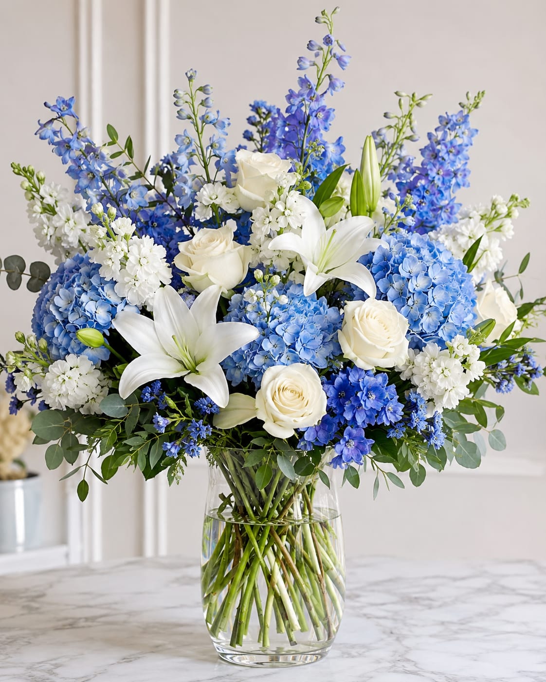 Clear Sky by Petal Perfection  - A cool-toned bouquet of blue hydrangeas and white lilies that feels like a breath of fresh air-ideal as a birthday bouquet, sympathy arrangement, or to mark an anniversary where you want to convey calm and elegance. The rounded design pairs full, cloud-like blue hydrangeas with crisp white roses and lilies, while tall blue stock-like spires add graceful height and movement so the recipient feels both soothed and celebrated.  Arranged in a clear glass vase with visible stems for a modern, clean presentation, this composition blends blue, white, and green for a refined, airy look. The mix of textures-from the hydrangeas' softness to the lilies' sculptural petals-creates visual interest up close and from across the room. Choose this arrangement when you want the person you're thinking of to feel comforted, uplifted, and truly seen; sizing and delivery options are available at checkout.